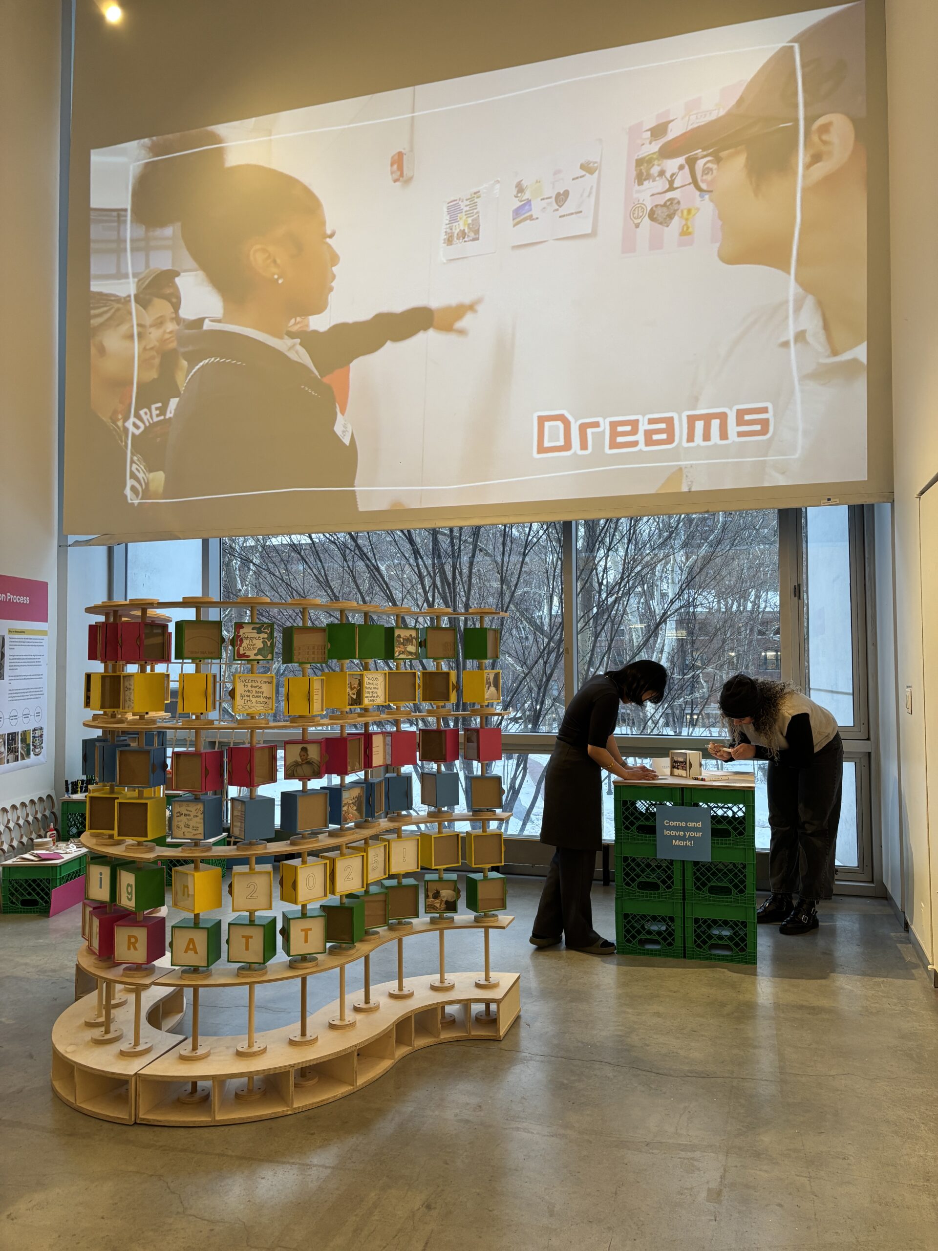 Exhibition gallery interior with a large projected video on the wall showing people in conversation, including on-screen text reading “Dreams.” In the foreground, a colorful, multi-tiered sculptural installation made of small cube-like modules spells out “Pratt 2025.” Two visitors stand at a green crate-based table labeled “Come and leave your mark,” writing or interacting with materials. Large windows behind the installation reveal leafless trees outside, indicating a daytime winter setting.