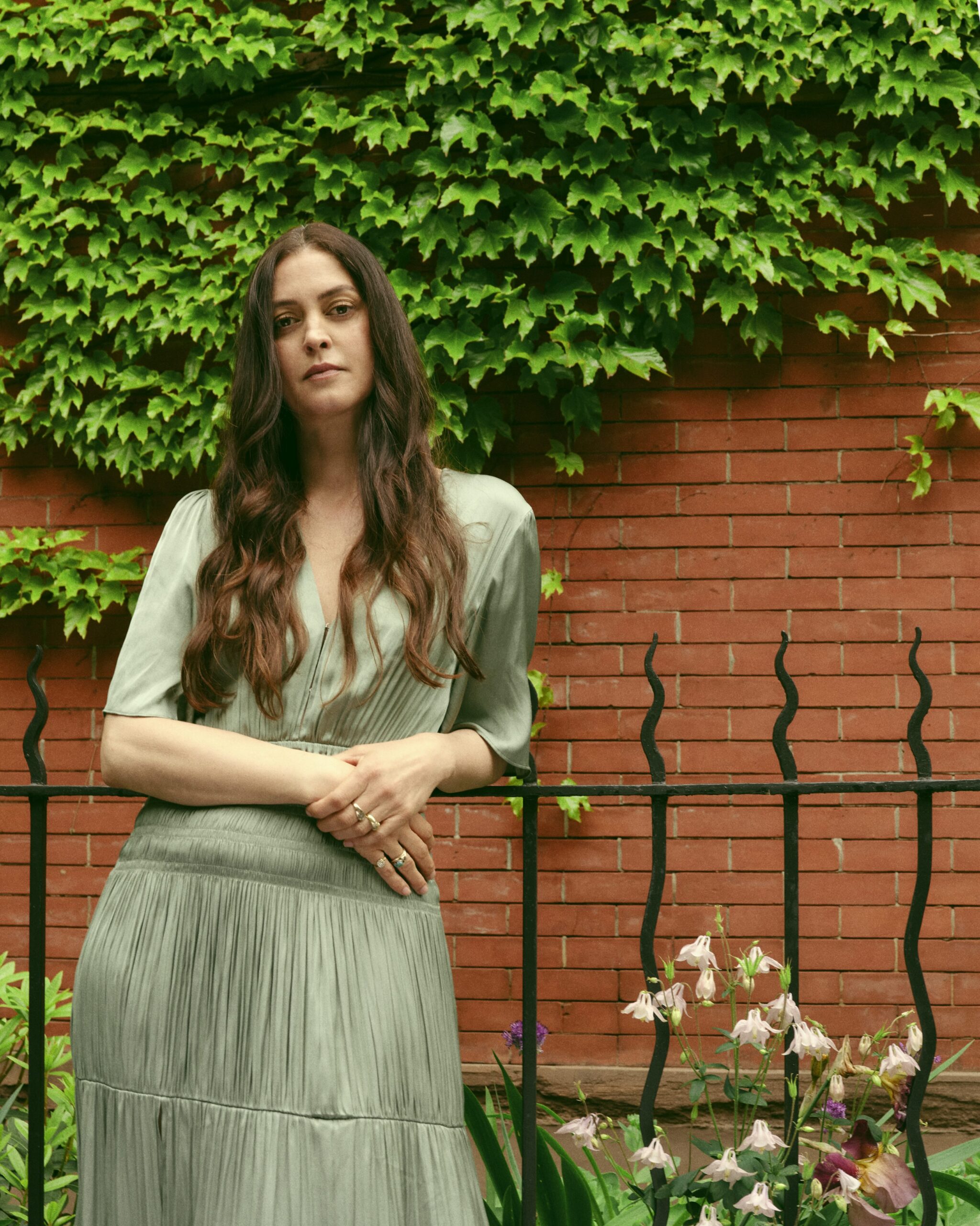 Portrait of a woman standing outdoors in front of a red brick wall covered with green ivy. She wears a light sage-green, short-sleeved dress with a pleated skirt and rests her hands together at her waist. A black metal fence runs across the foreground, with flowering plants and greenery at the base. The subject looks directly at the camera with a neutral expression in a garden-like setting.