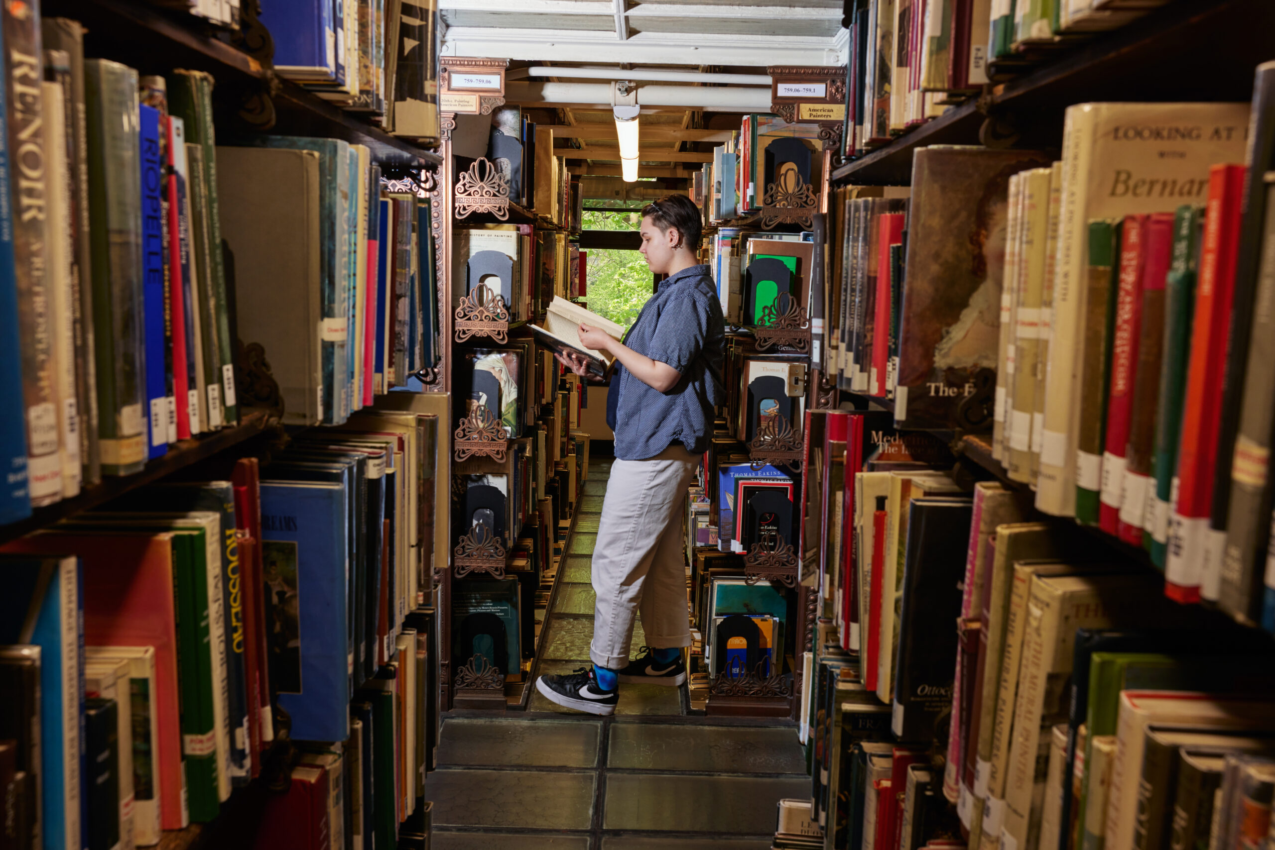 A person stands between two tall shelves filled with books in a library. They are holding an open book, appearing to read, with a focused expression. The shelves are lined with a variety of colorful book covers. A window in the background lets in natural light, illuminating the space. The person is wearing a short-sleeved blue shirt and light-colored pants.