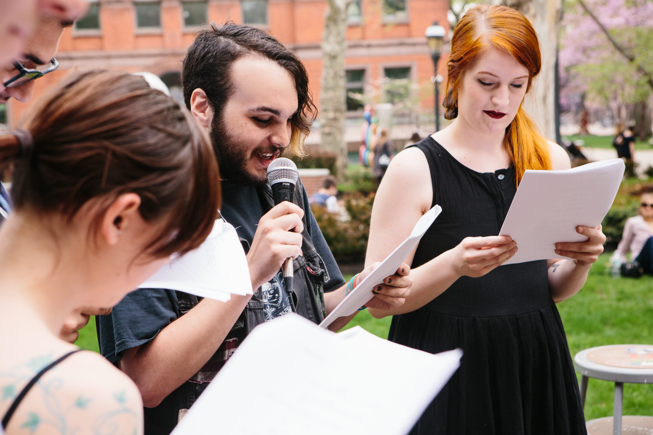 Outdoor gathering where several people read from printed pages; one person speaks into a handheld microphone while another stands nearby reading, with a campus lawn and brick building in the background.