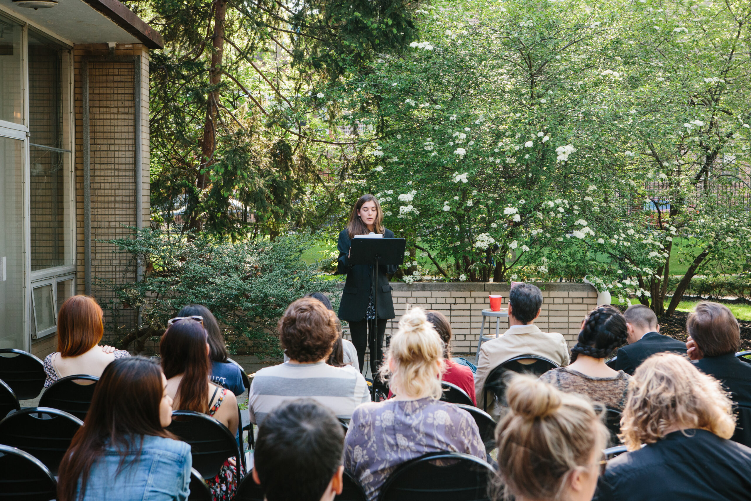 Outdoor reading or presentation taking place in a garden courtyard. A person stands at a lectern reading from papers while seated audience members face forward on folding chairs. The setting includes a brick building facade, leafy trees, and flowering shrubs, indicating a