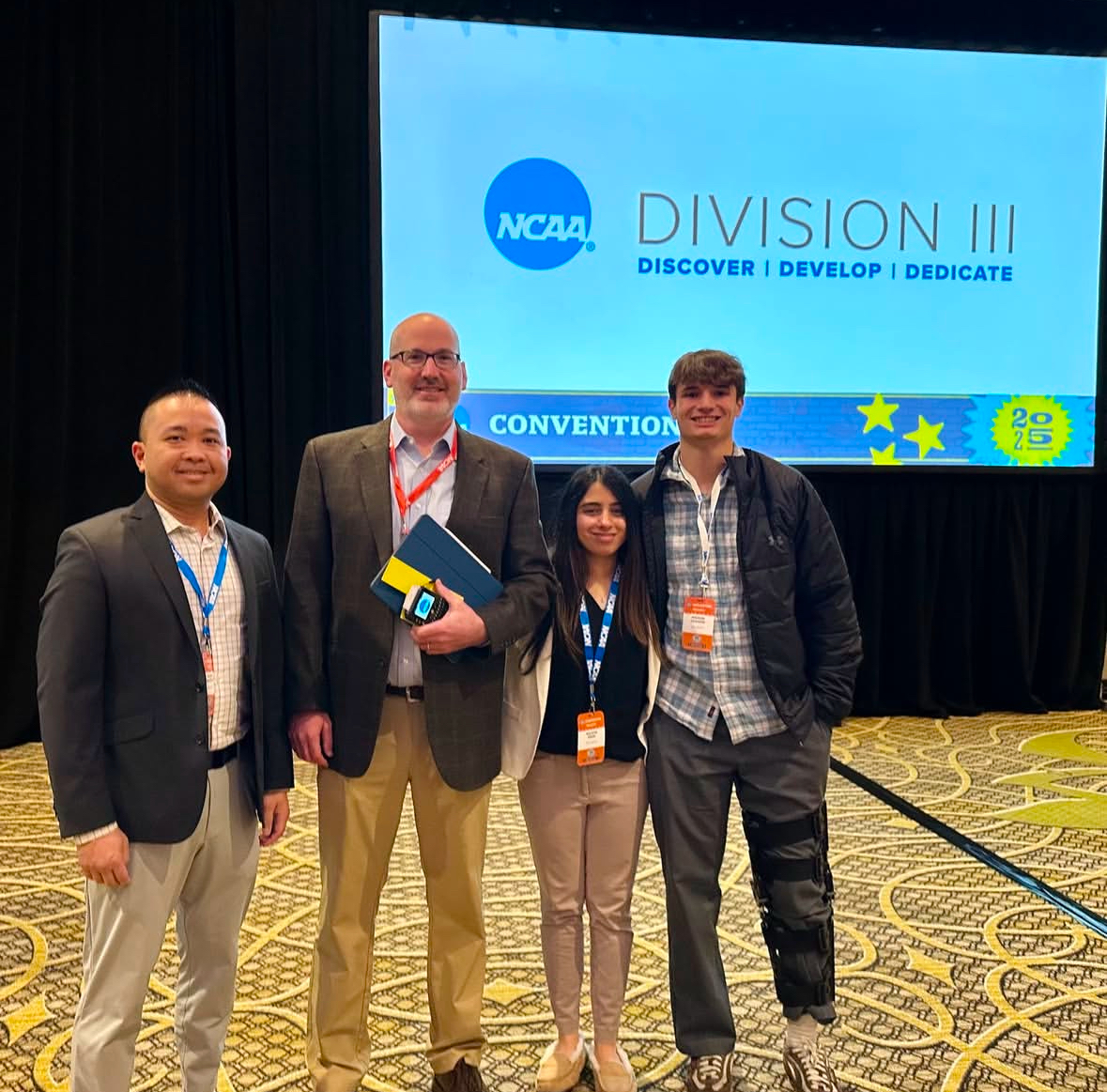 Four individuals pose in front of a screen displaying the NCAA Division III logo along with the text "Discover | Develop | Dedicate". They are smiling and standing closely together. The first person on the left wears a gray suit with a patterned shirt. The second person is in a tan jacket holding a notebook. The third person, dressed in light pants and a black top, stands in between them. The fourth person, wearing a jacket and a leg brace, stands on the far right. The background shows a dark curtain and a patterned carpet.