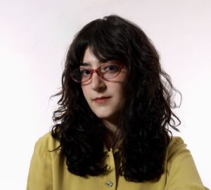 Studio head-and-shoulders portrait of a person with long dark wavy hair wearing red eyeglasses and a mustard-yellow blouse, looking slightly to the side against a light background.
