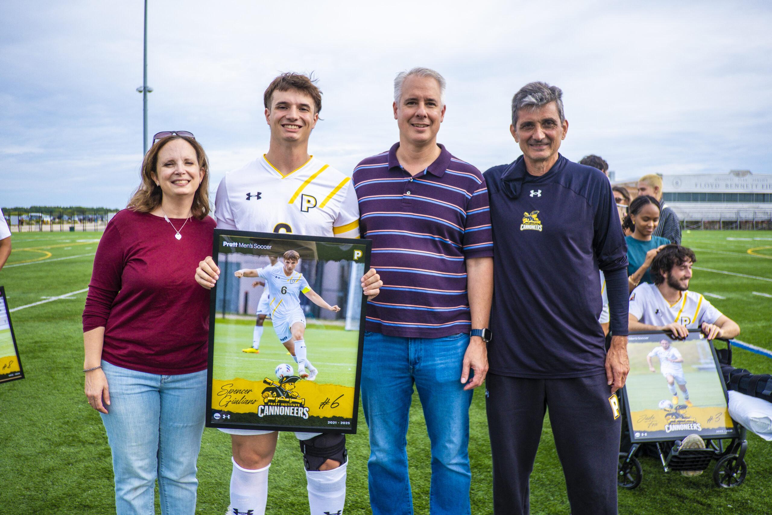 A young man in a white soccer jersey with yellow details poses with three adults on a sports field. He holds a poster of himself in action on the soccer field titled "Pratt Men's Soccer." The group is smiling, with a cloudy sky in the background and a soccer field visible. Other individuals are present in the background, also holding posters.