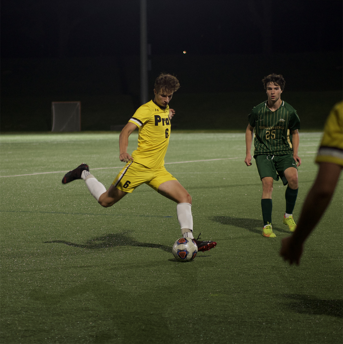 A soccer player in a yellow uniform with the number 6 is mid-kick, striking a soccer ball on a well-lit, dark green field. Another player in a green uniform with the number 25 watches from a short distance. The scene captures the action of an evening soccer match.