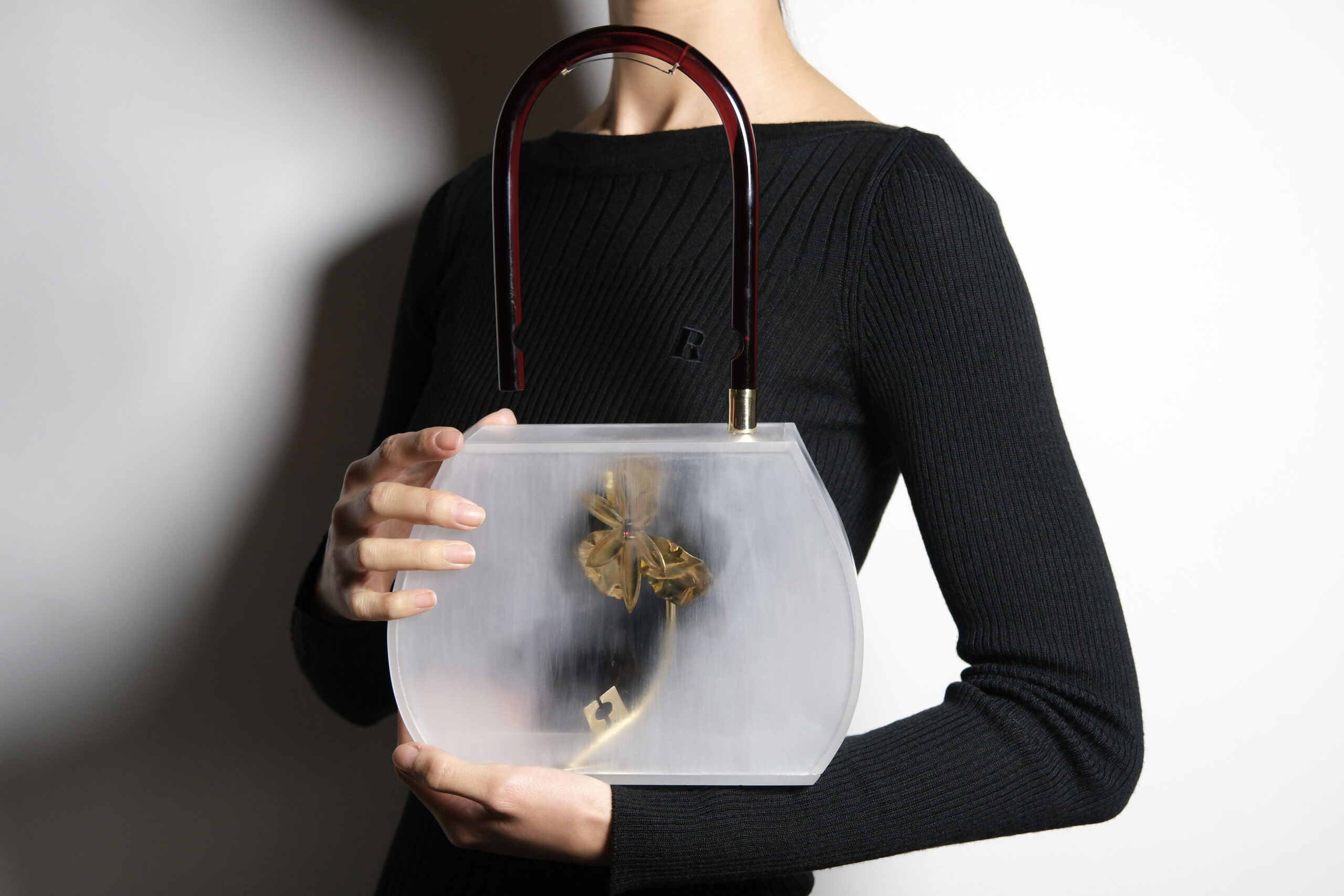 A person is holding a stylish, transparent handbag with a curved shape. The bag features a reddish-brown handle and contains a delicate, golden flower inside. The individual is wearing a black, ribbed long-sleeve top, and the background is a soft light gradient.