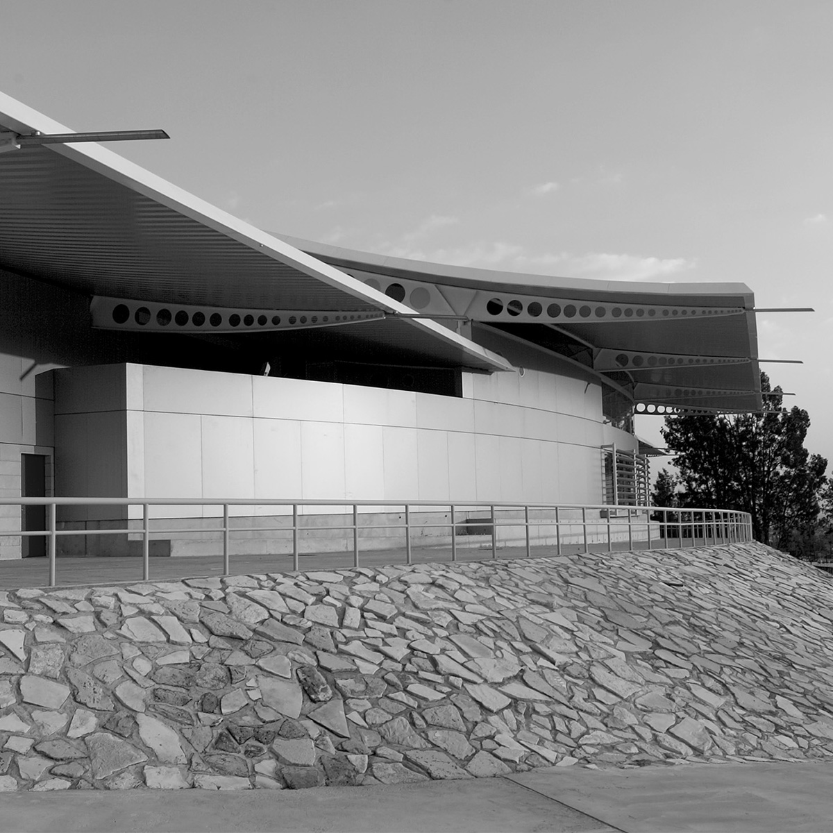 Black-and-white photograph of a modernist building with a curved roof and extended cantilevered canopy. The structure features circular cutouts along the roof edge and smooth, light-colored exterior walls. A metal railing runs along a walkway in the foreground, which is elevated above a stone retaining wall composed of irregularly shaped rocks. Trees and open sky appear in the background.