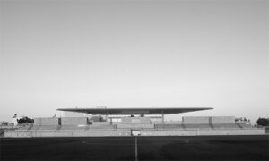 Black-and-white panoramic photograph of a low-profile modern building with a long, flat roof and horizontal emphasis. The structure sits behind a broad, open paved or grassy foreground, creating strong linear separation between ground and building. The sky is clear and expansive, with minimal surrounding elements, emphasizing the building’s silhouette and scale.