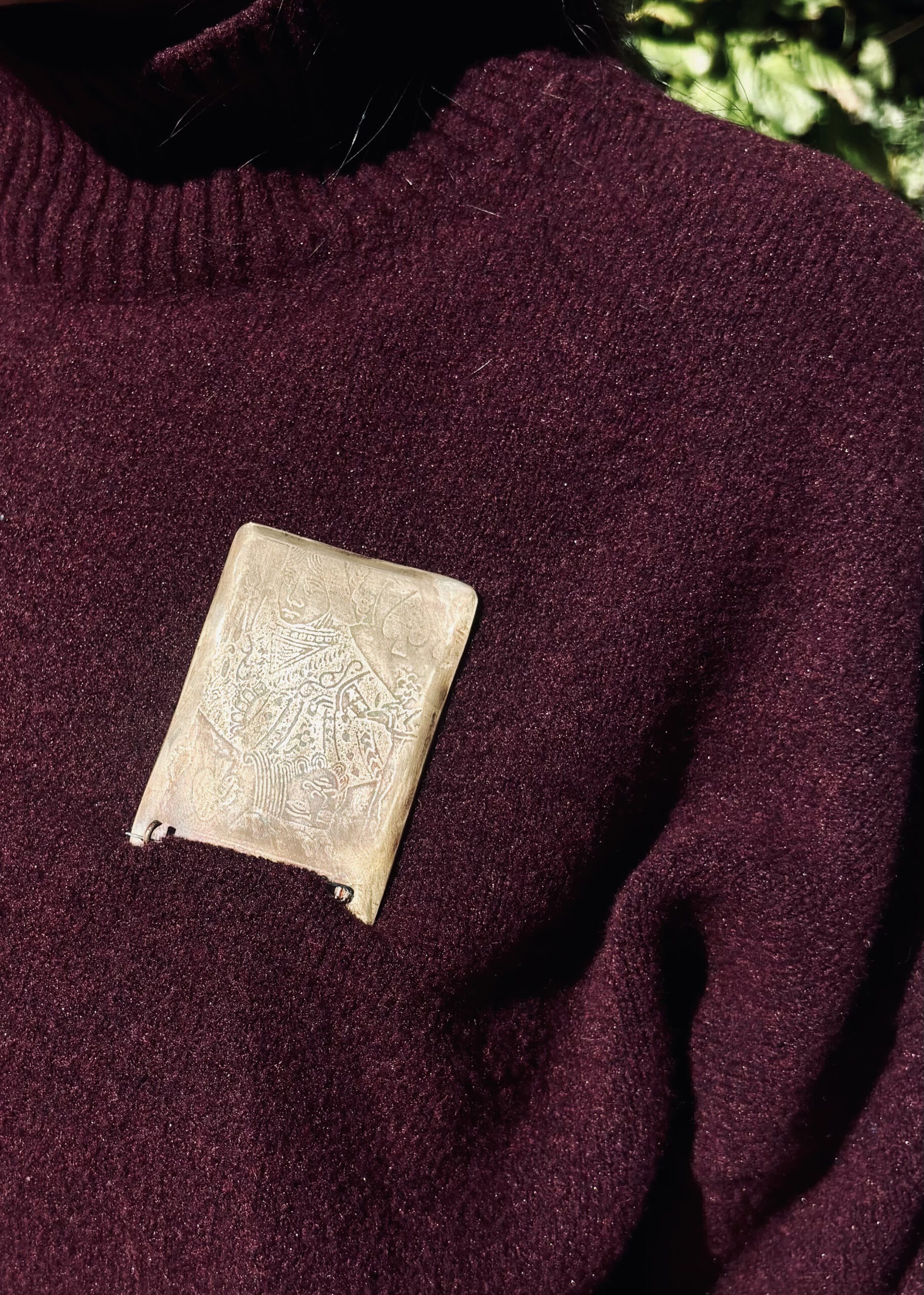 A close-up of a maroon sweater with a metallic brooch attached to it. The brooch has an intricate embossed design featuring figures and patterns. Sunlight highlights the texture of the fabric and the metallic surface, creating a sophisticated contrast.