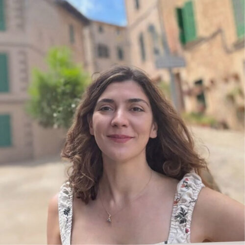 Portrait of a woman outdoors in a courtyard with stone buildings and green shutters in the background. She faces the camera with a slight smile. Her hair is loose and shoulder-length, and she wears a sleeveless patterned top and a small pendant necklace. The background is softly blurred.
