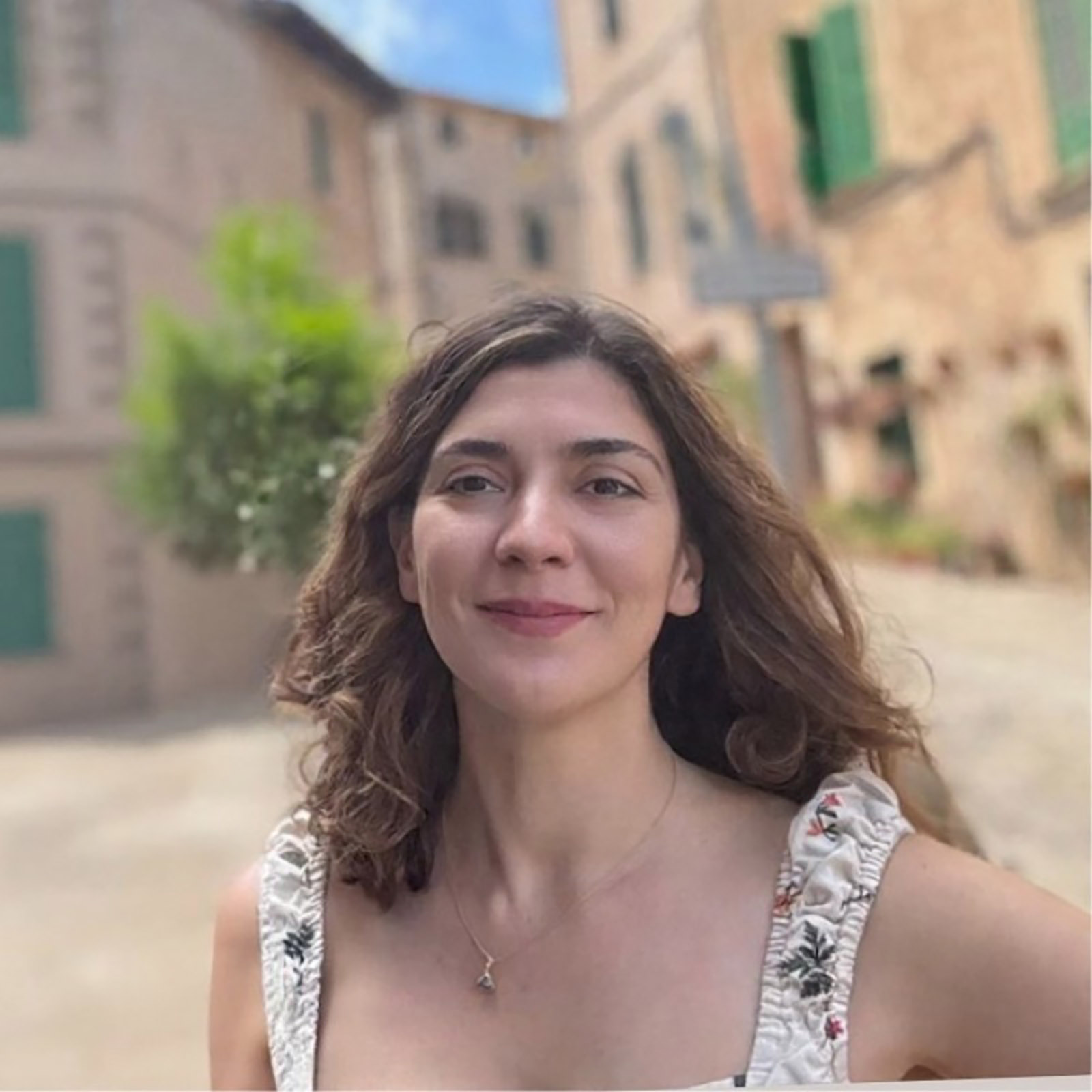 Portrait of a woman outdoors in a courtyard with stone buildings and green shutters in the background. She faces the camera with a slight smile. Her hair is loose and shoulder-length, and she wears a sleeveless patterned top and a small pendant necklace. The background is softly blurred.