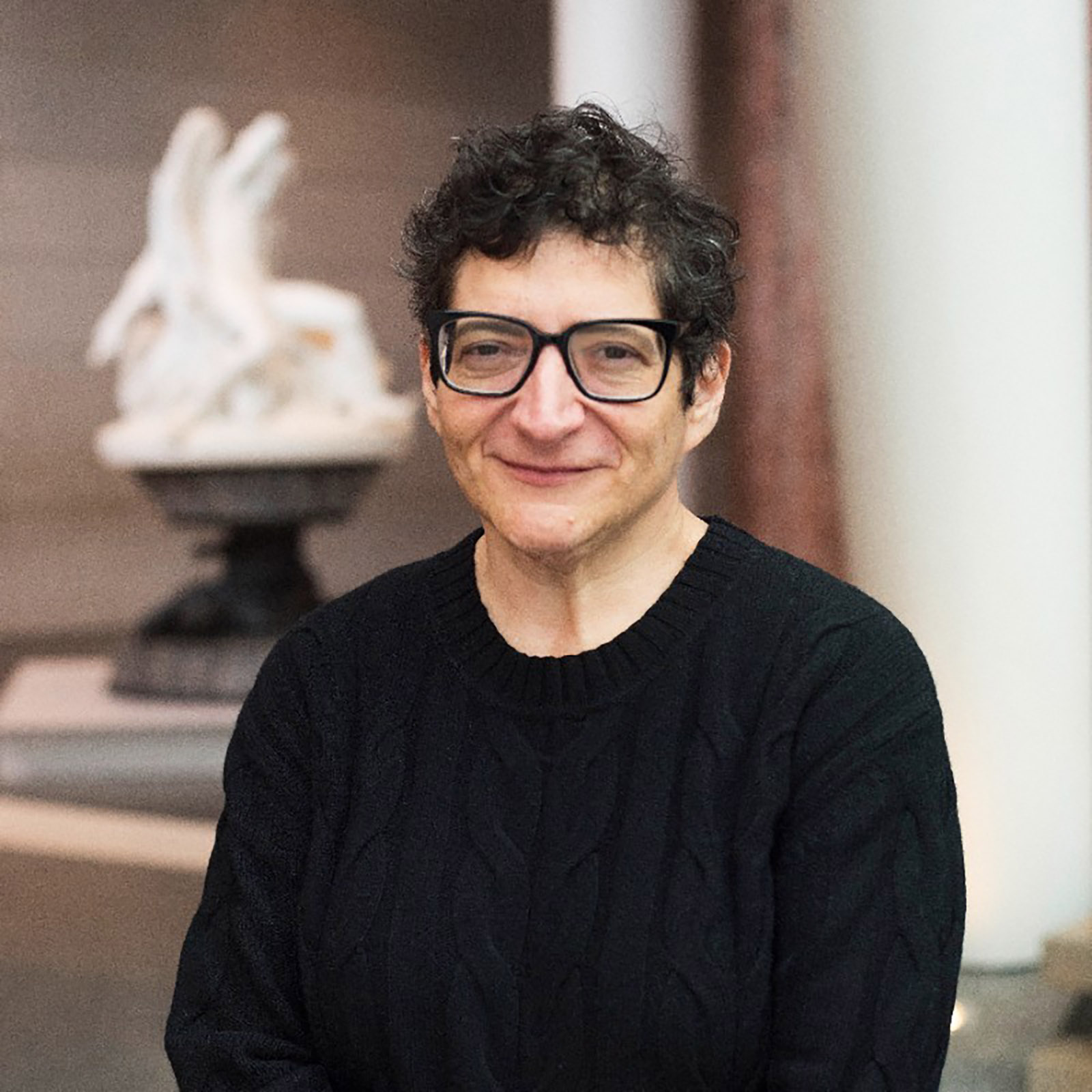 Portrait of a person wearing glasses and a dark sweater, standing indoors. The person smiles slightly while facing the camera. A softly blurred sculpture and interior architectural elements appear in the background.