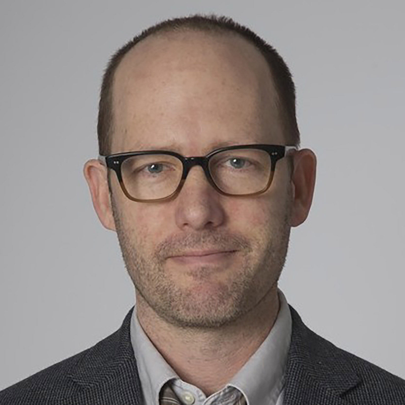Headshot of a person wearing glasses, a collared shirt, tie, and textured jacket against a plain light background. The person faces forward with a neutral expression. The image is cropped closely around the head and shoulders.