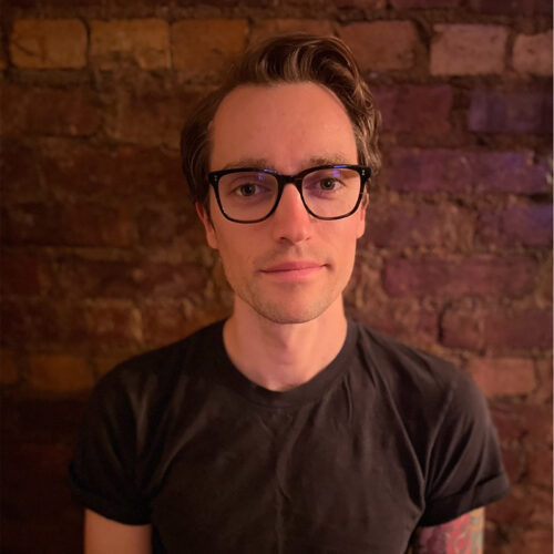 Head-and-shoulders portrait of a person wearing dark-framed glasses and a black T-shirt, standing in front of a brick wall. The person looks directly at the camera with a neutral expression. Warm indoor lighting casts soft shadows on the brick background.