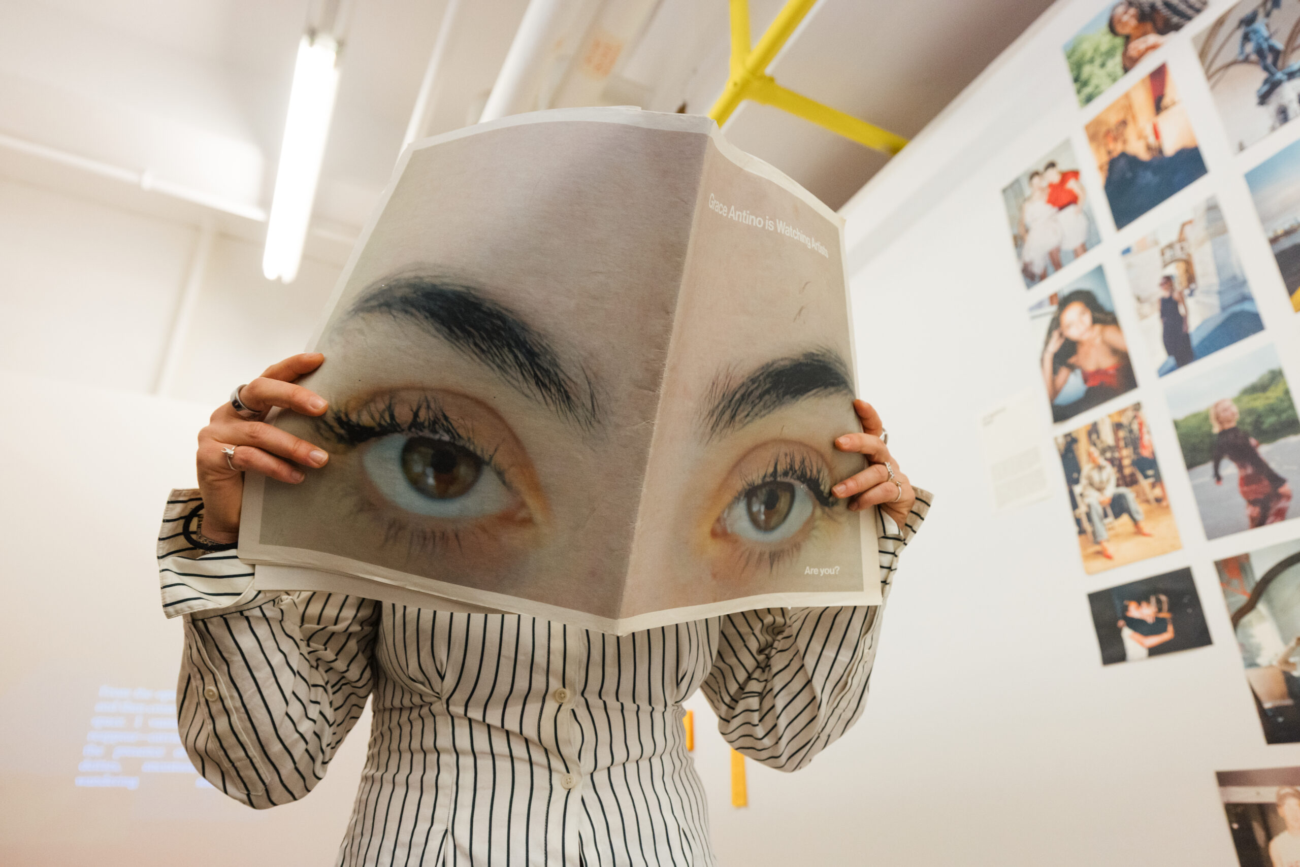 A person holds a large magazine page featuring close-up images of eyes, obscuring their face. They wear a striped shirt and have rings on their fingers. In the background, a wall is decorated with various photos. The setting appears to be an art exhibition or gallery.