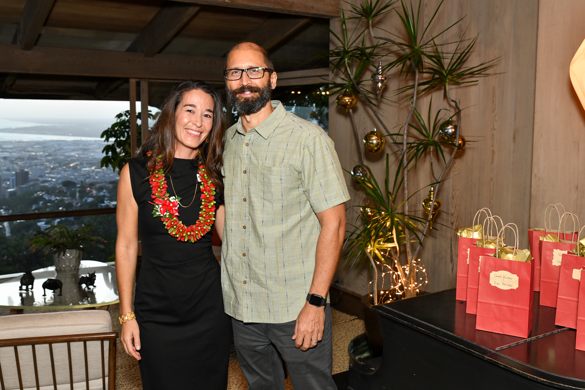 The same woman, Kristi Roberson Cardoso, is dressed in a black sleeveless dress with a floral lei, standing next to a man, Fabio Cardoso, with a beard, wearing a light green short-sleeve shirt, both smiling in a well-lit indoor setting.