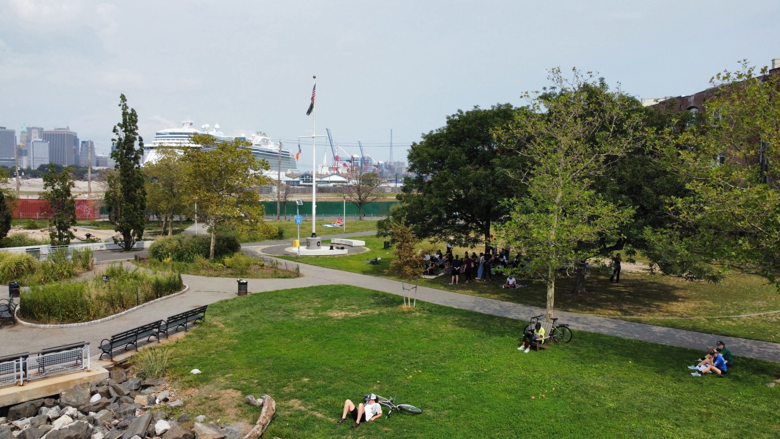 A panoramic view of a park featuring green grass, trees, and walking paths. In the foreground, several people are relaxing, some on the ground or sitting on benches. In the background, a large cruise ship is docked, with the city skyline visible on the left side. A few individuals are gathered together in a shaded area, enjoying a social gathering.