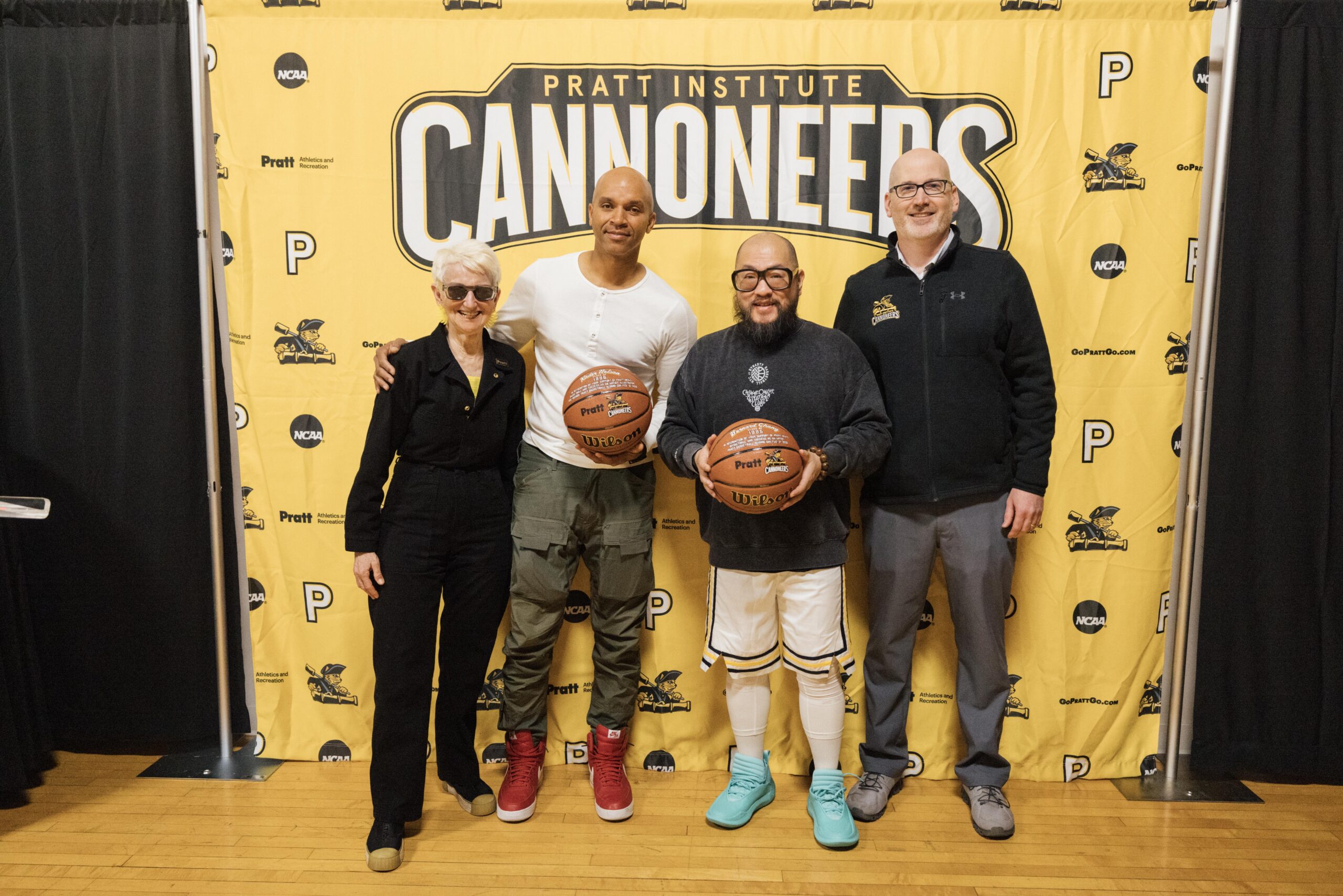 Four people pose together in front of a large yellow backdrop featuring the Pratt Institute Cannoneers logo. The group includes a woman in a black outfit and sunglasses, a man in a white shirt and green cargo pants, a man in a black sweater and shorts, and a man in a black jacket. Each person holds a basketball, and the setting appears to be a celebratory event related to the institute.