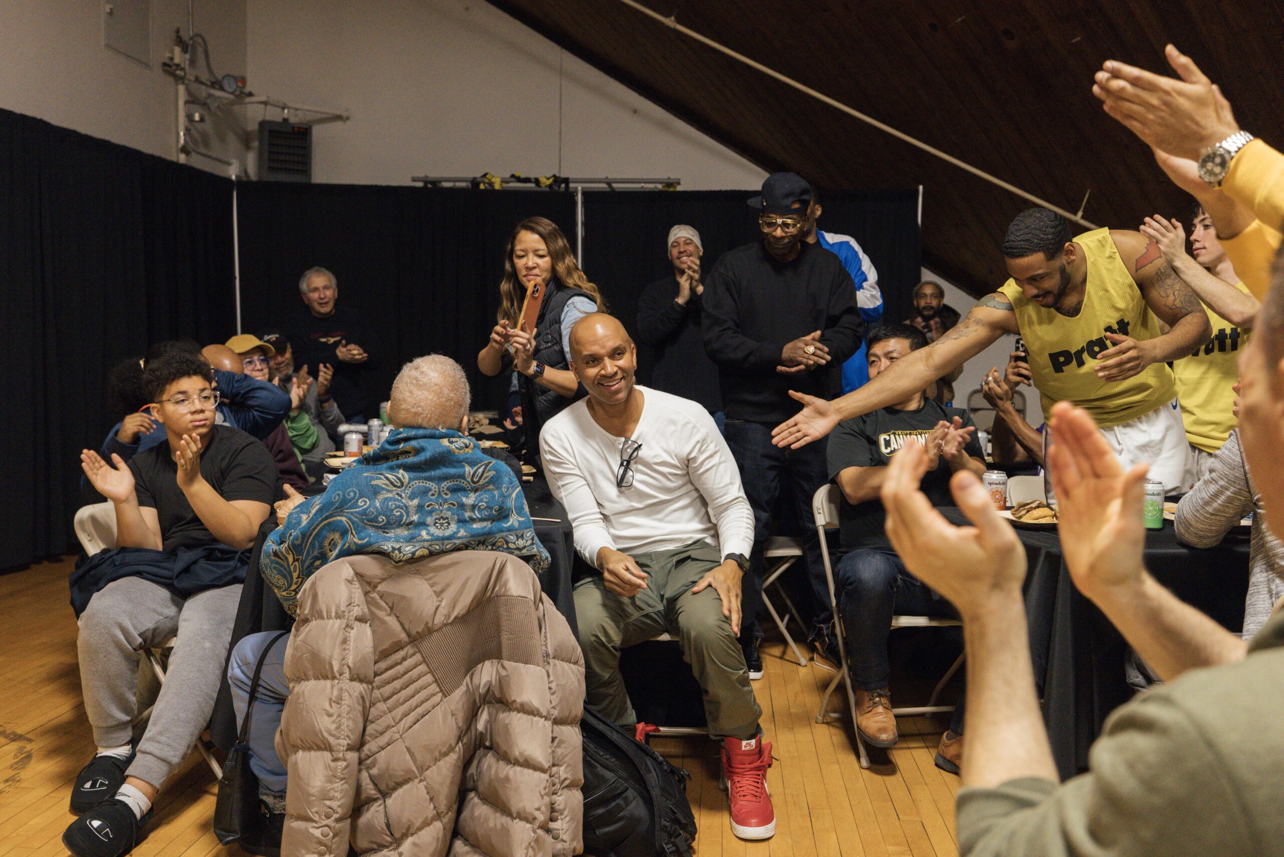 A group of people is gathered indoors, clapping and celebrating. In the foreground, a smiling man in a white shirt and red shoes sits on a chair, while others around him, including a woman with a flute, join in applause. Some individuals are seated at tables in the background, and the atmosphere appears joyful and lively, with a mix of ages present. A black curtain is visible behind them, adding to the indoor setting.