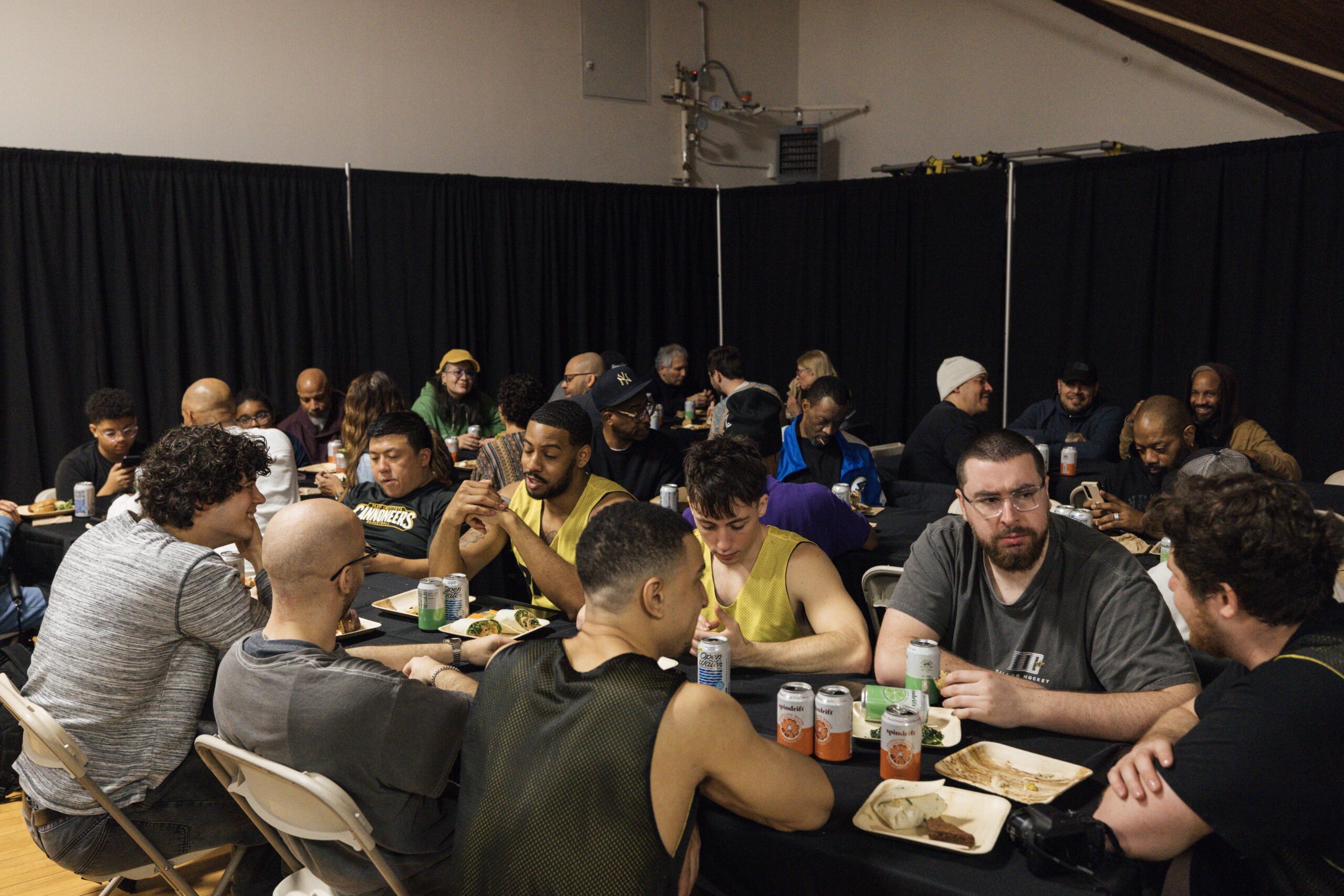 A large group of people is seated around tables in a dining area, sharing a meal. Many individuals are engaged in conversation, with some looking at their phones. Several participants are wearing sports jerseys or casual attire. There are plates of food and drinks on the tables, while a backdrop of black curtains creates a cozy atmosphere. The lighting is warm, highlighting the diverse group of attendees.