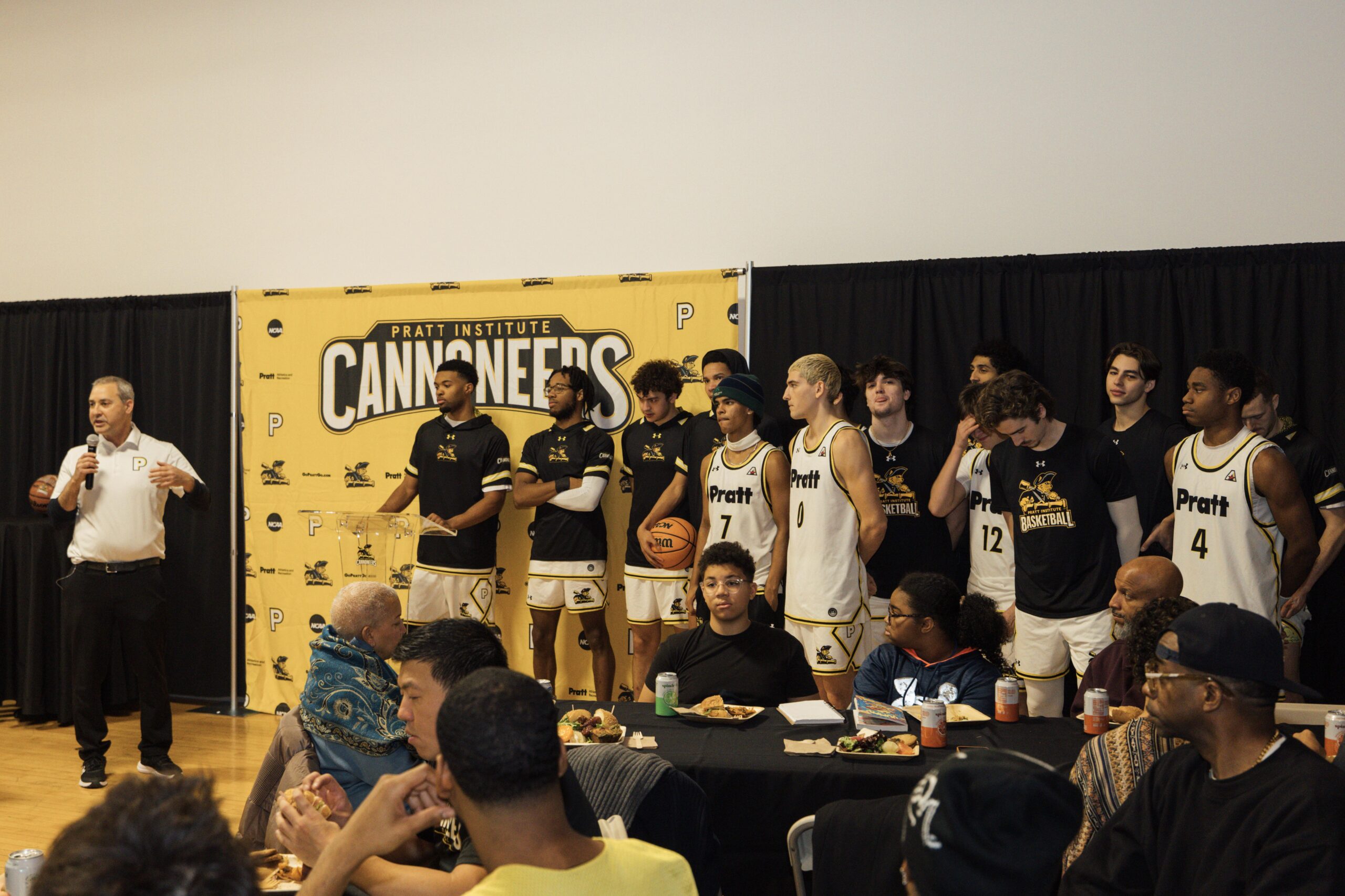 A group of male basketball players stands on a stage with a yellow Pratt Institute Cannoneers banner behind them. One man is speaking into a microphone, while the players in uniforms listen. In the foreground, several attendees are seated at a table, enjoying food and drinks. The atmosphere appears to be a team event or gathering, with a focus on team spirit and community.