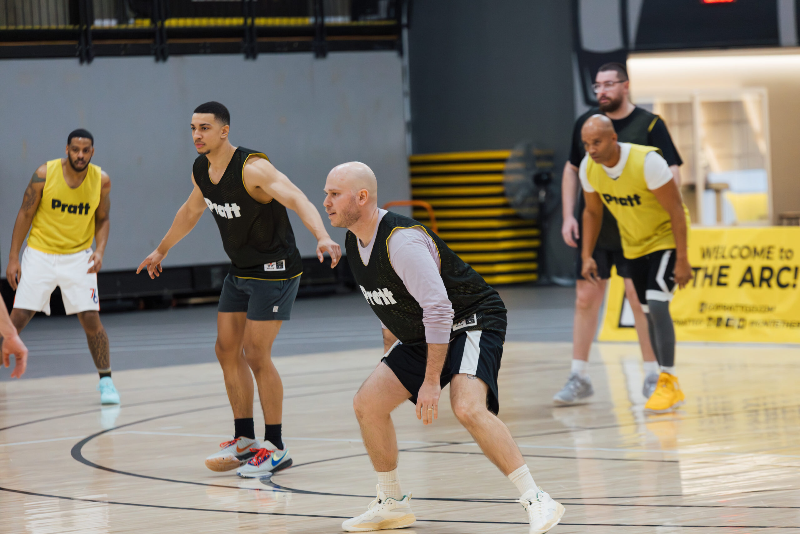 A group of five men playing basketball in a gym. Two players are wearing yellow jerseys with “Pratt” printed on them, while three players are in black jerseys. They are in active stances on the court, focused on the game. The background features bleachers and a sign welcoming visitors to "THE ARC." The gym has a polished wooden floor and modern lighting.