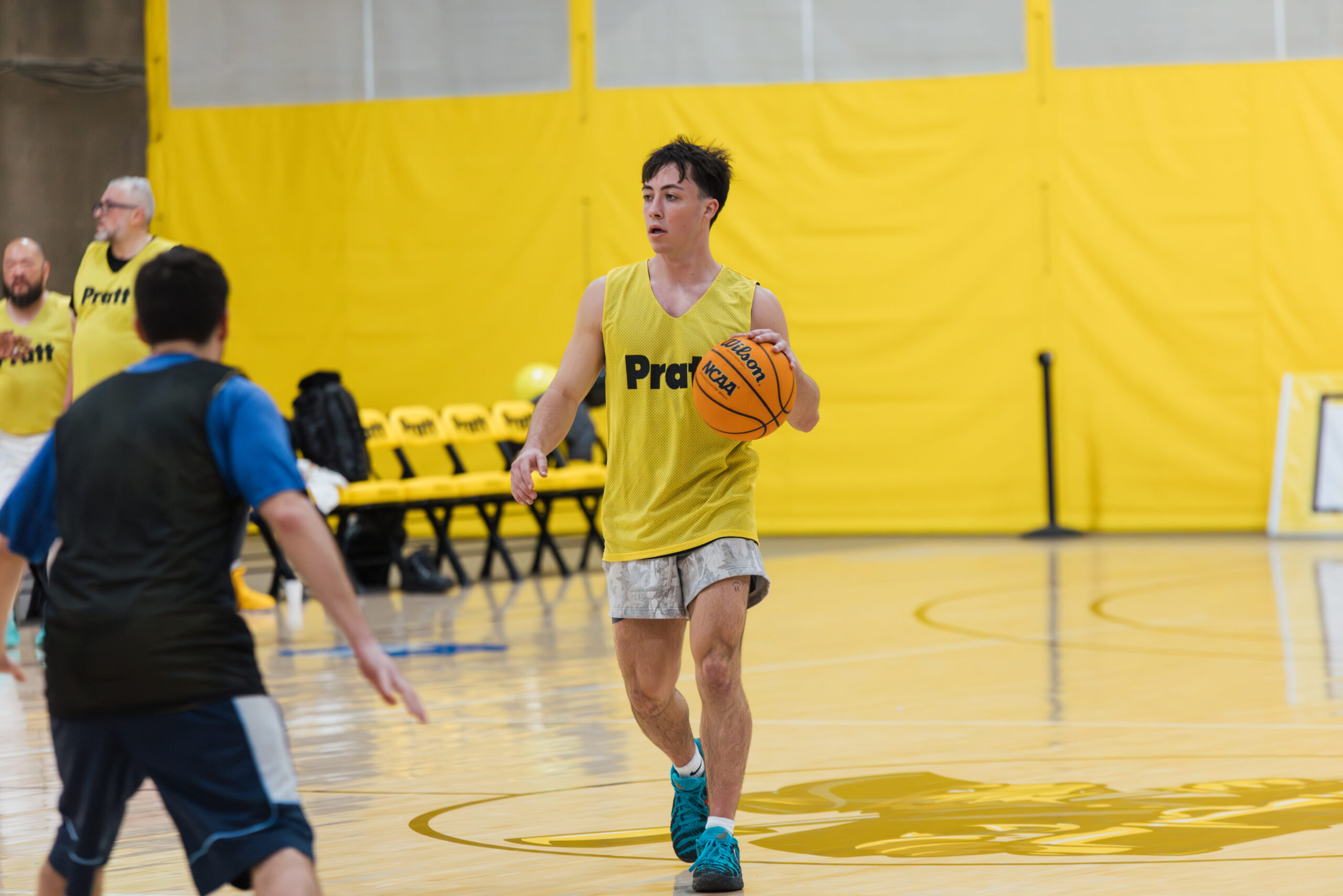 A player in a yellow practice jersey dribbles a basketball on a wooden gym floor, with a bright yellow background and empty bleachers. Another player, dressed in a blue shirt, is partially visible in the foreground. The atmosphere is energetic, suggesting a basketball practice or game setting.