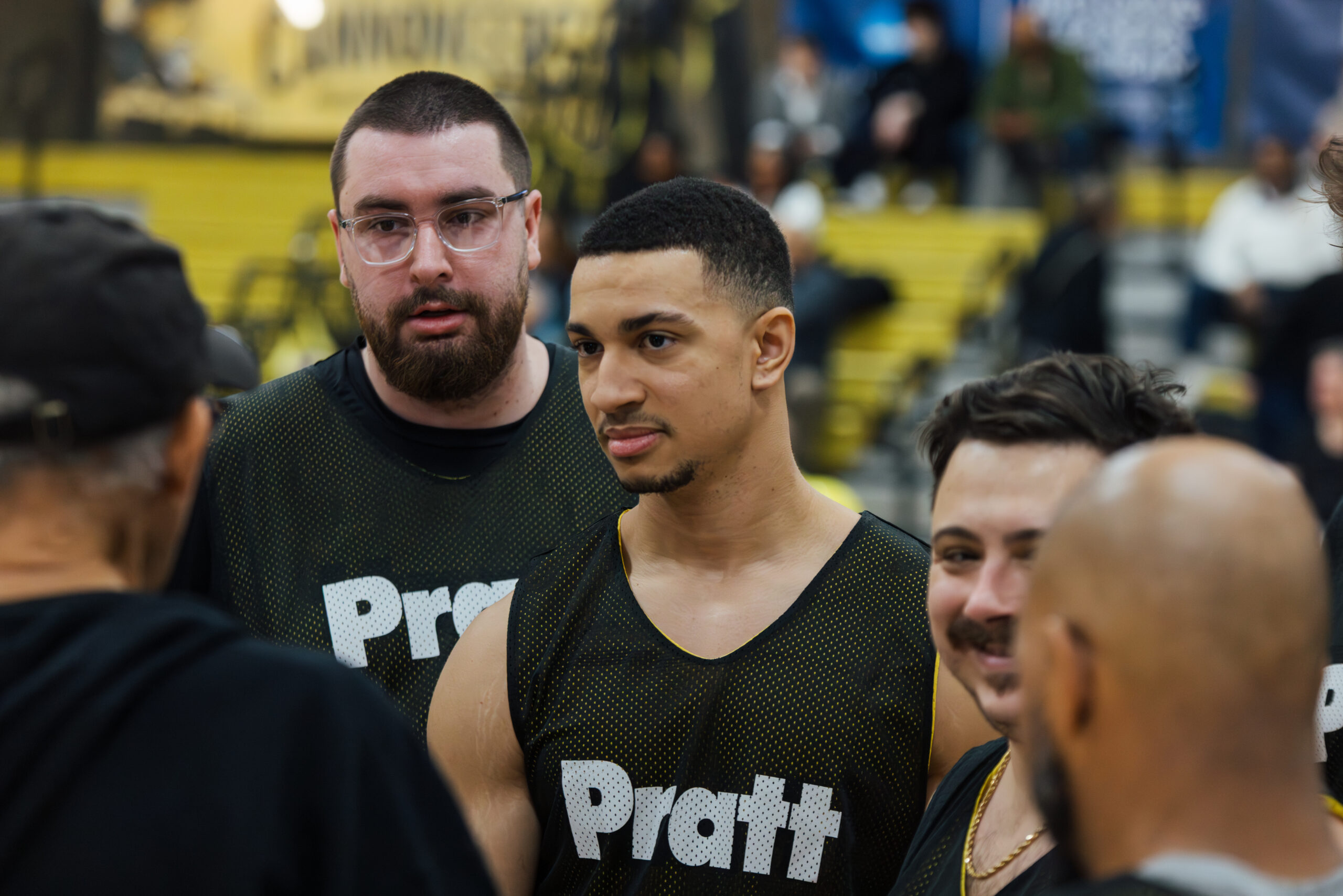 A group of four basketball players are gathered in a gym, wearing black jerseys with the word "Pratt" printed on them. One player stands in the foreground, looking attentively at someone off-camera. Two teammates beside him are engaged in conversation, with one wearing glasses and a beard and the other having a light mustache. The gym has a colorful backdrop and a crowd in the stands.