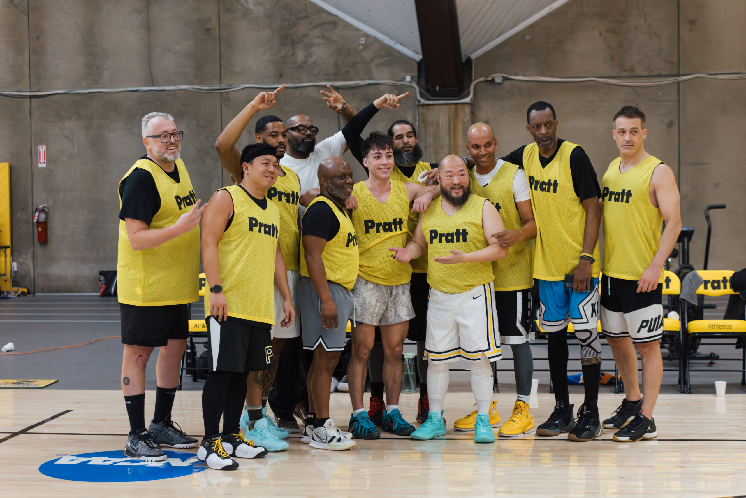 A group of basketball players stands together in a gym, all wearing yellow jerseys with "Pratt" printed on them. They are smiling and posing for the camera, with some giving thumbs up and others pointing. The background features basketball equipment and seating. There are a mix of players in terms of body type and ethnicity, and they seem to be enjoying a team moment.