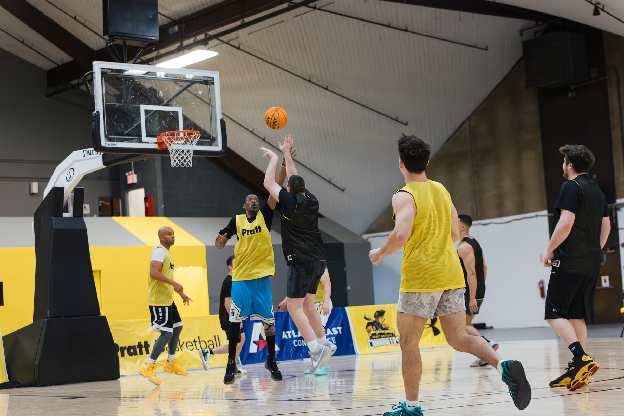 A basketball game in an indoor gym features several players in yellow and black jerseys. One player is shooting the ball towards the hoop while another defends. The court has a bright wooden floor, and banners are displayed along the walls. The setting is lively with players actively engaged in the game.