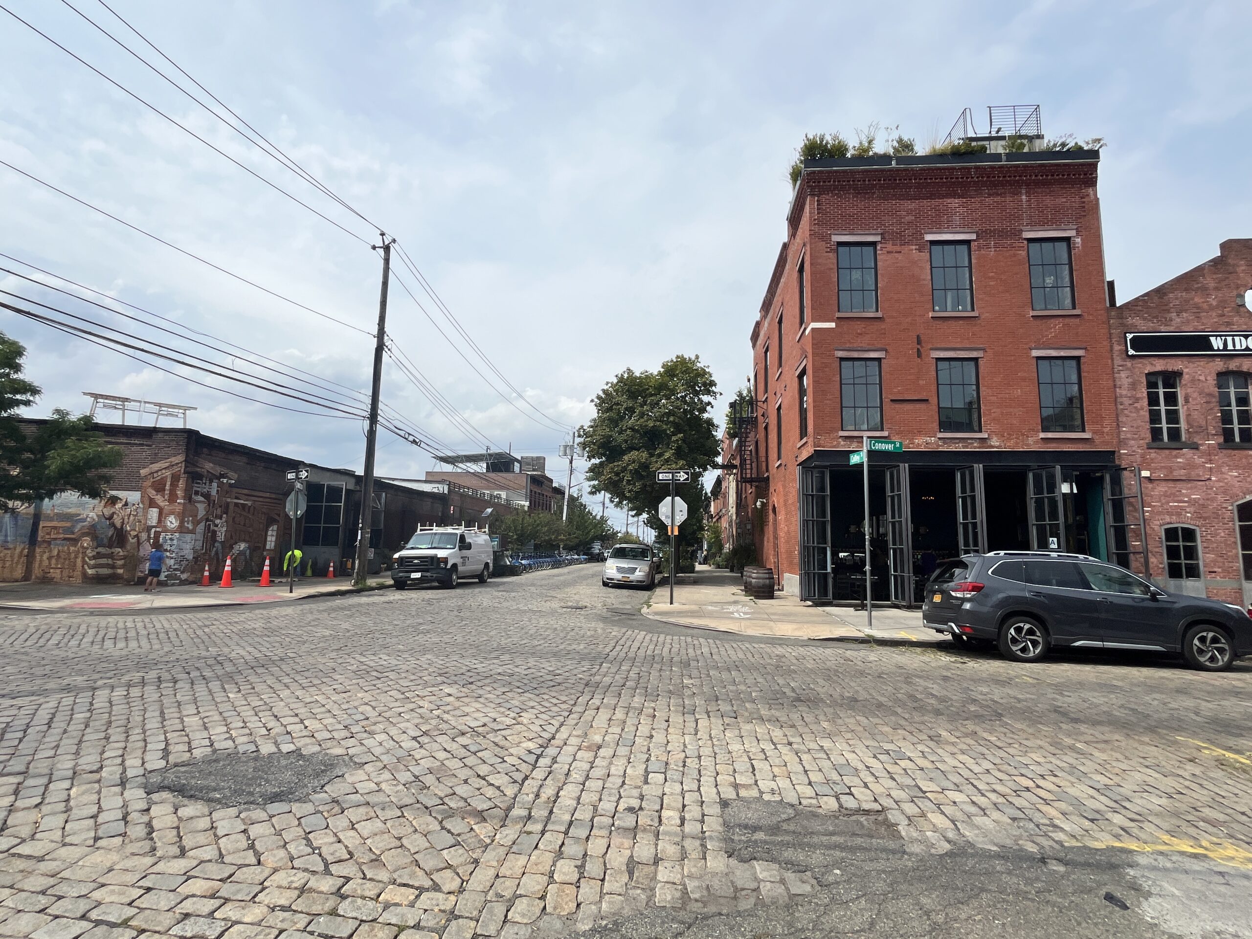 A cobblestone street intersects at a corner with a red brick building featuring large windows. A white truck and a dark SUV are parked nearby. Traffic cones line the sidewalk, and a few people are walking along the street. In the background, there's a mural on an adjacent building and additional structures visible. The sky is partly cloudy.