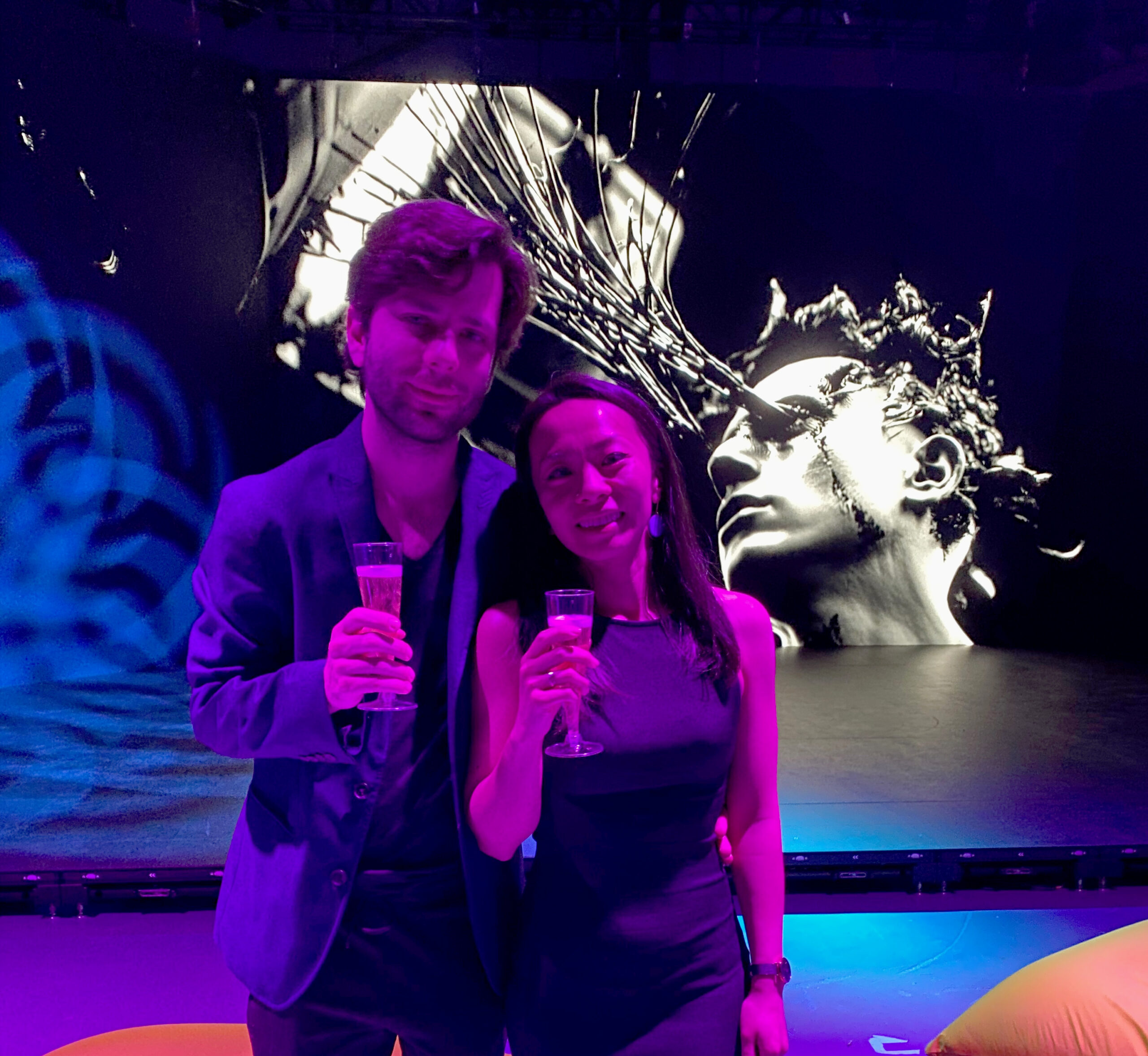 a man, Alex Iselin, and a woman, Jane Wu, pose together holding glasses of champagne, with a colorful abstract background.