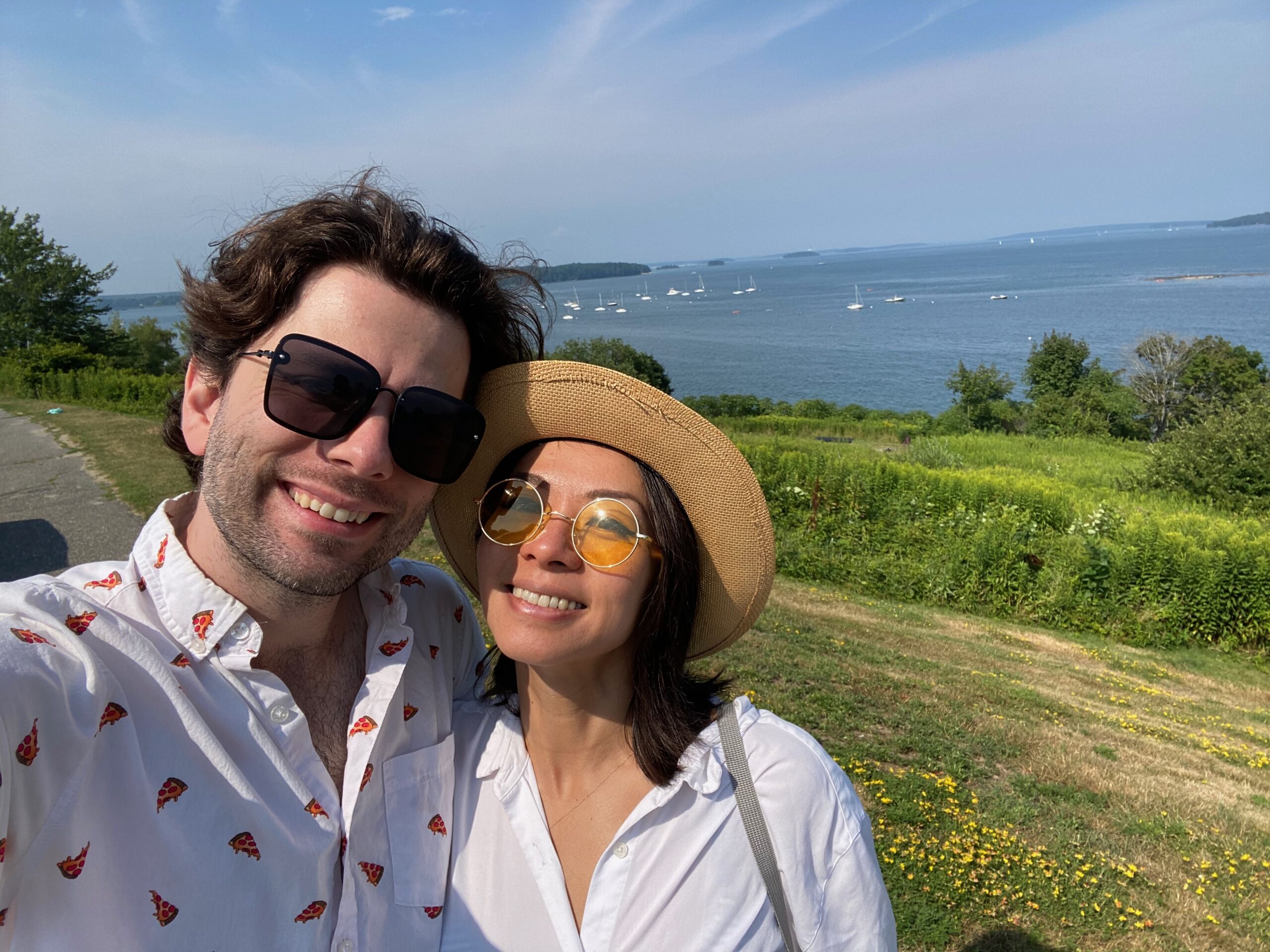 A couple, Alex Iselin and Jane Wu, takes a selfie outdoors near a scenic waterfront, smiling against a backdrop of greenery and distant boats on the water. Both individuals wear sunglasses; the woman has a straw hat.