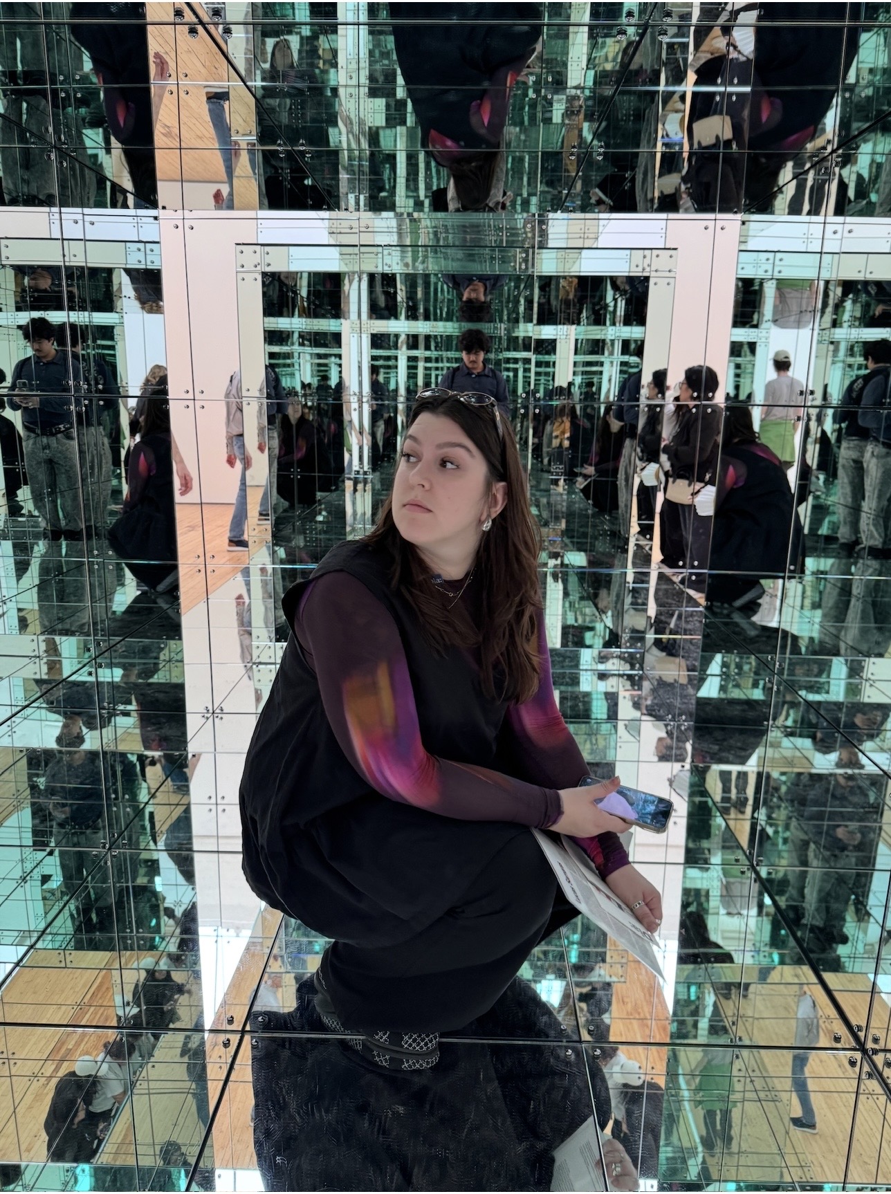 A person with long brown hair is sitting on a black surface in a mirrored installation. They are wearing a black outfit with a colorful long-sleeve top and holding a phone in one hand and a piece of paper in the other. The environment is filled with reflections from multiple mirrored surfaces, creating an infinite effect. In the background, several other visitors can be seen interacting with the installation.