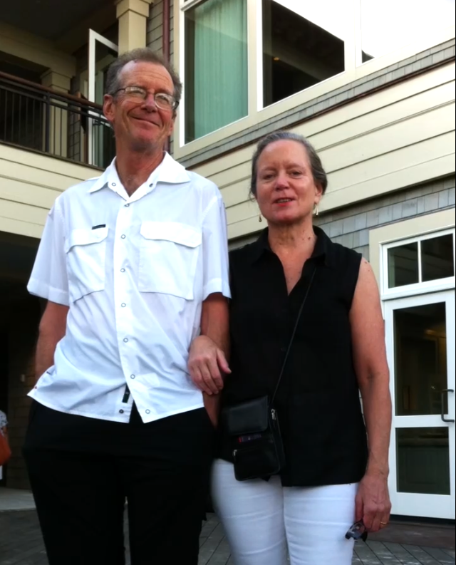 A color photo depicts an elderly man, Michael Thompson, and woman, Jan Eagers, standing together outside a modern building, both smiling and dressed casually. The background shows windows and wood siding.