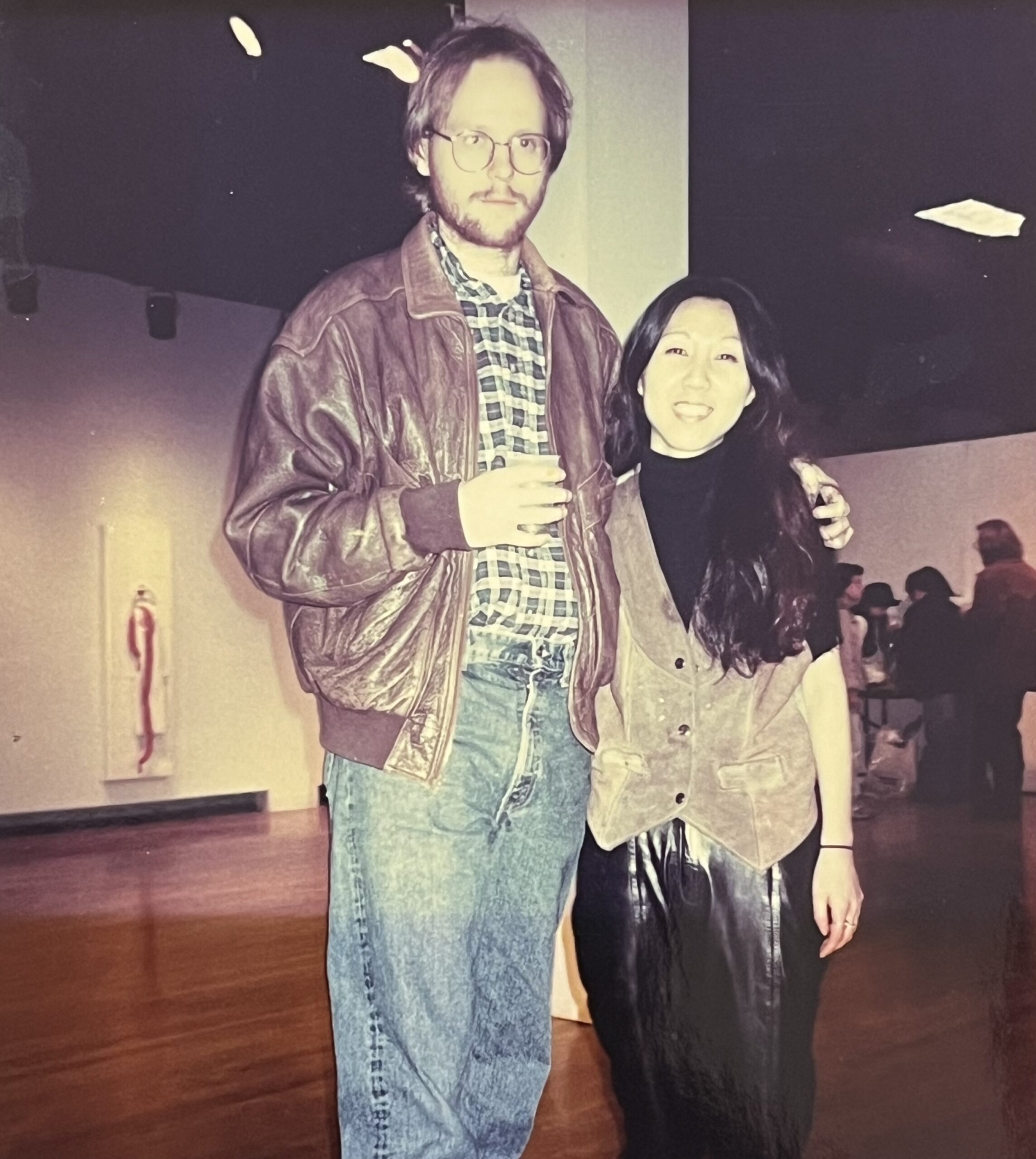 A man, Jeff Gabel, with glasses and a brown leather jacket stands beside a woman, Jaye Moon, with long black hair, both smiling at a social event.