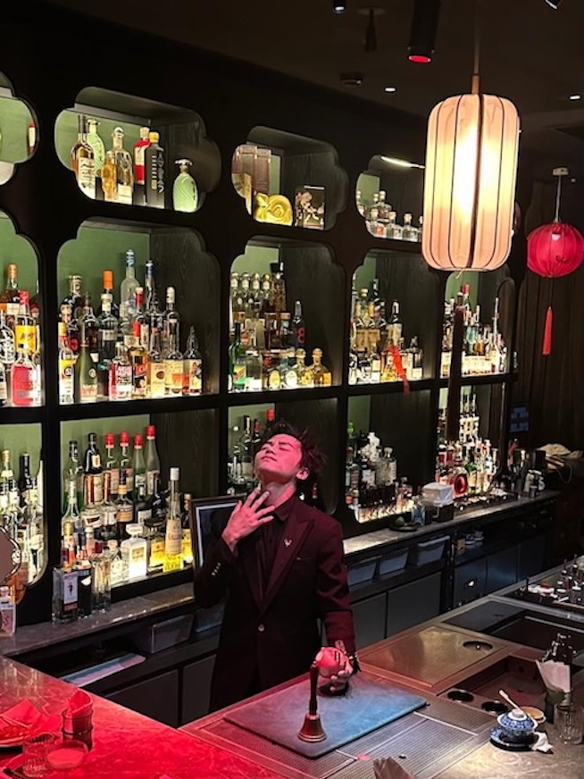 A dimly lit bar interior with a unique ceiling made of hanging glass bottles. Tables with candles are scattered throughout, and patrons are engaged in conversation. A bartender in a dark suit is posing confidently, with a variety of liquor bottles displayed behind him on shelves, illuminated by soft lighting. The bar area has a red glow and includes a cocktail preparation space.