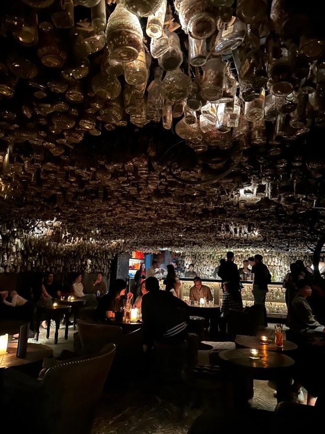 A dimly lit bar interior with a unique ceiling made of hanging glass bottles. Tables with candles are scattered throughout, and patrons are engaged in conversation.