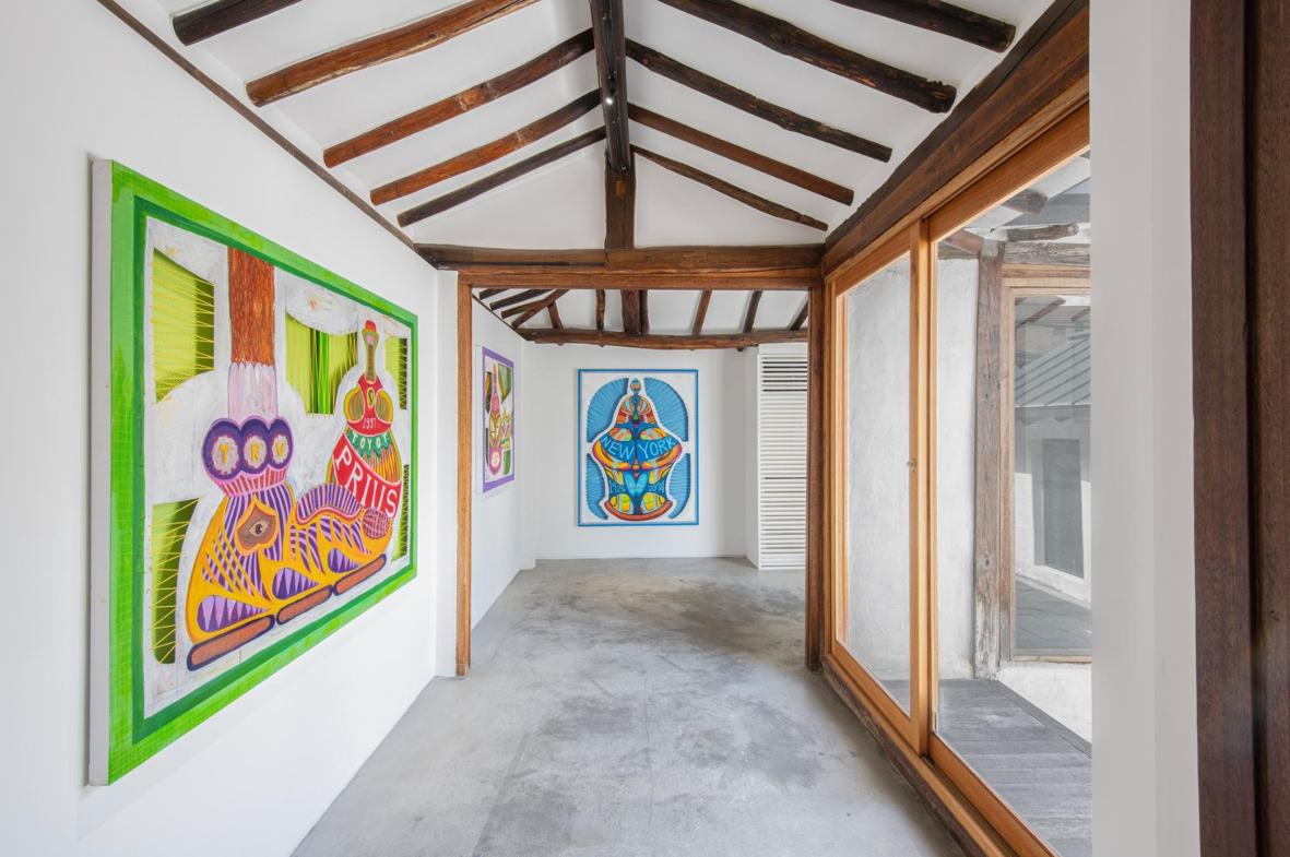 A brightly lit gallery space featuring two colorful art pieces on white walls. One artwork has green and purple elements with a design labeled "Prius," while the other features a vivid blue background with a central design labeled "NEW YORK." The ceiling is wooden with exposed beams, and there are large glass windows letting in natural light. The floor is plain concrete.