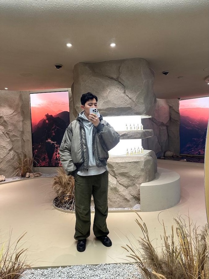 Lee stands indoors taking a selfie in front of a rock-themed display with screens showing mountain landscapes.