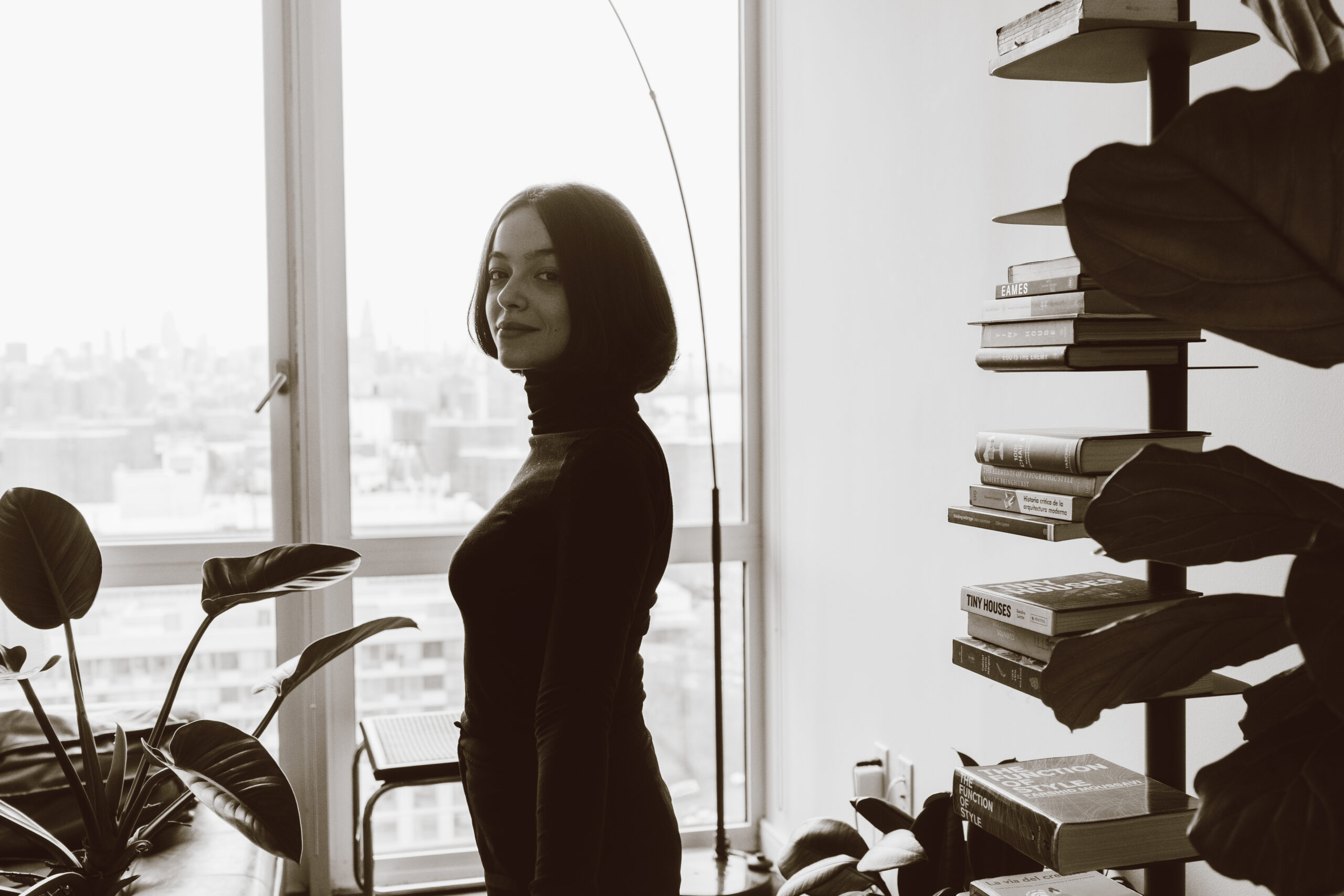 A woman with shoulder-length dark hair is positioned near a large window, looking slightly over her shoulder. She wears a black high-neck top. In the foreground, there is a plant with broad leaves, and to the right, a bookshelf filled with neatly arranged books. The scene is in black and white, with a city skyline visible in the background.