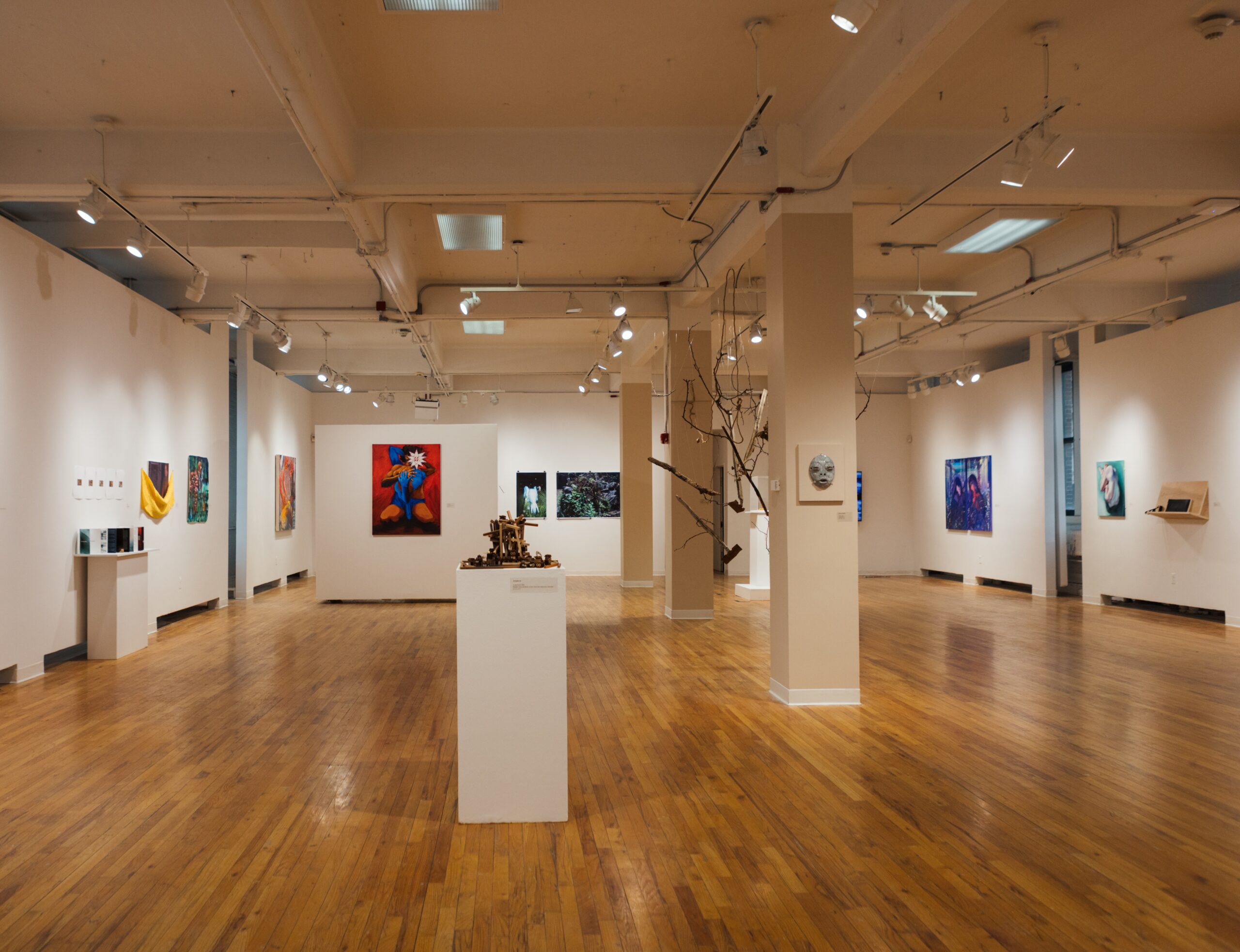 An art gallery featuring wooden flooring and white walls. Various artworks, including paintings in vibrant colors and textures, are displayed on the walls. A central pedestal showcases a sculptural piece made of natural materials, while several light fixtures illuminate the space. The atmosphere appears bright and open, encouraging viewers to explore the art installations.