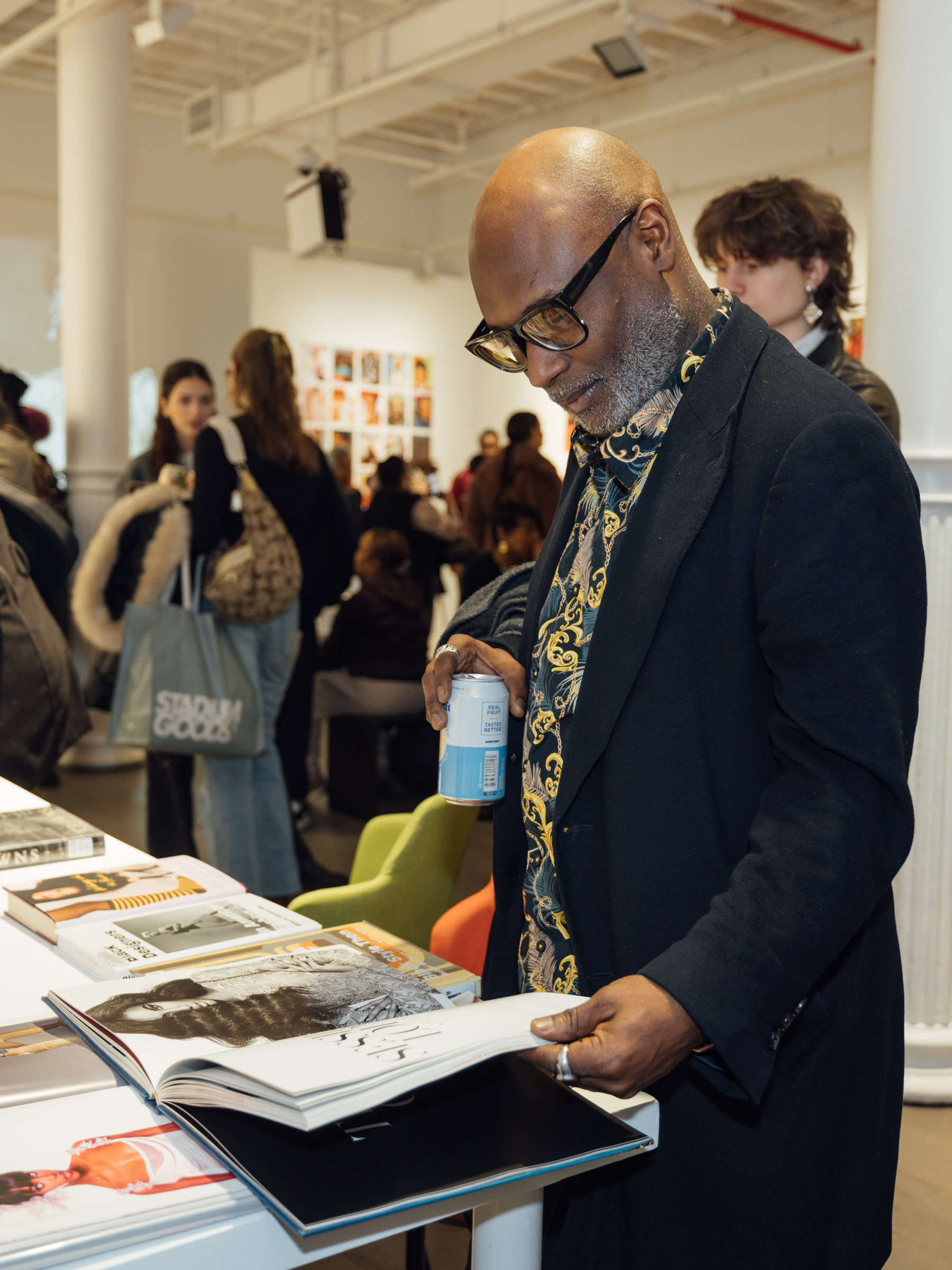 A man with a bald head and glasses is closely examining a magazine or book while standing at a table filled with various publications. He wears a black coat over a patterned shirt and holds a can in his other hand. In the background, a busy event or gathering is taking place with people mingling and sitting, and a wall decorated with photographs is visible.