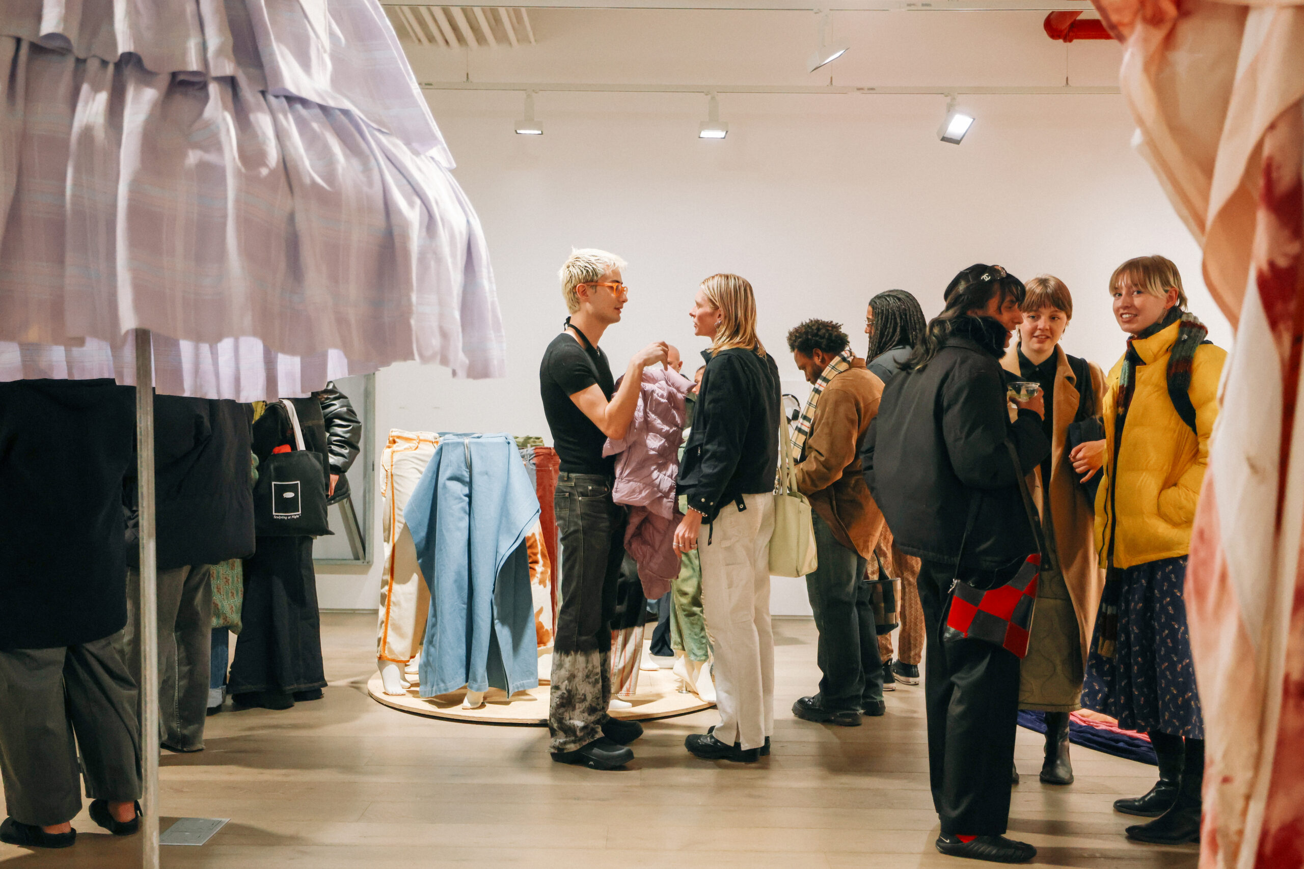 A group of people at a fashion exhibition are engaged in conversation and browsing clothing displayed on hanging racks. In the foreground, a man with light hair, wearing a black shirt, interacts with a woman holding a light purple garment. Other attendees in the background are scattered, chatting and examining various outfits, which include colorful denim and patterned fabrics. The room is well-lit with modern lighting.