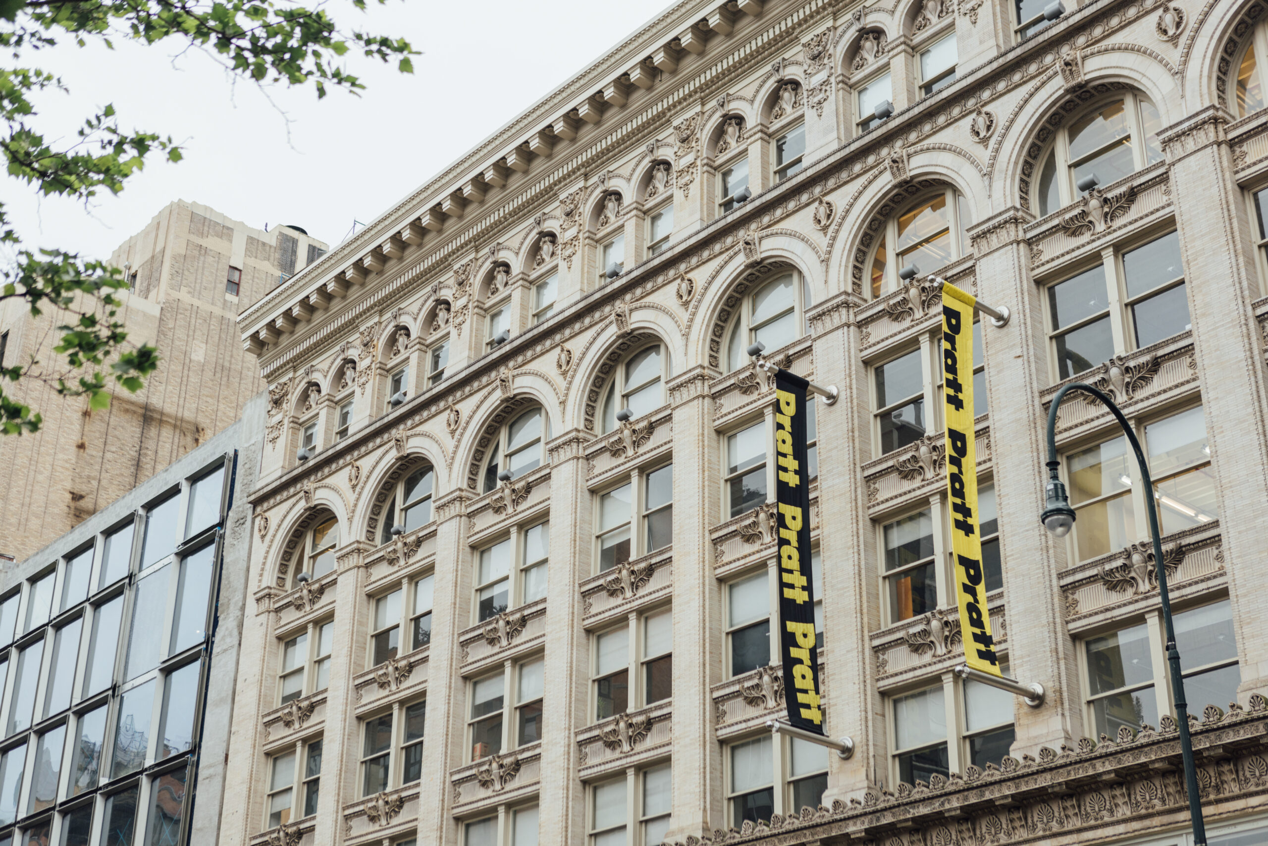 A close-up view of a historic building's façade, showcasing intricate architectural details such as arches and ornamental designs. Two large black banners with yellow text displaying the word "Pratt" are hanging from the building. The photo also features a modern glass structure beside the historical building, framed by green foliage in the foreground. The sky appears overcast, contributing to the urban atmosphere.