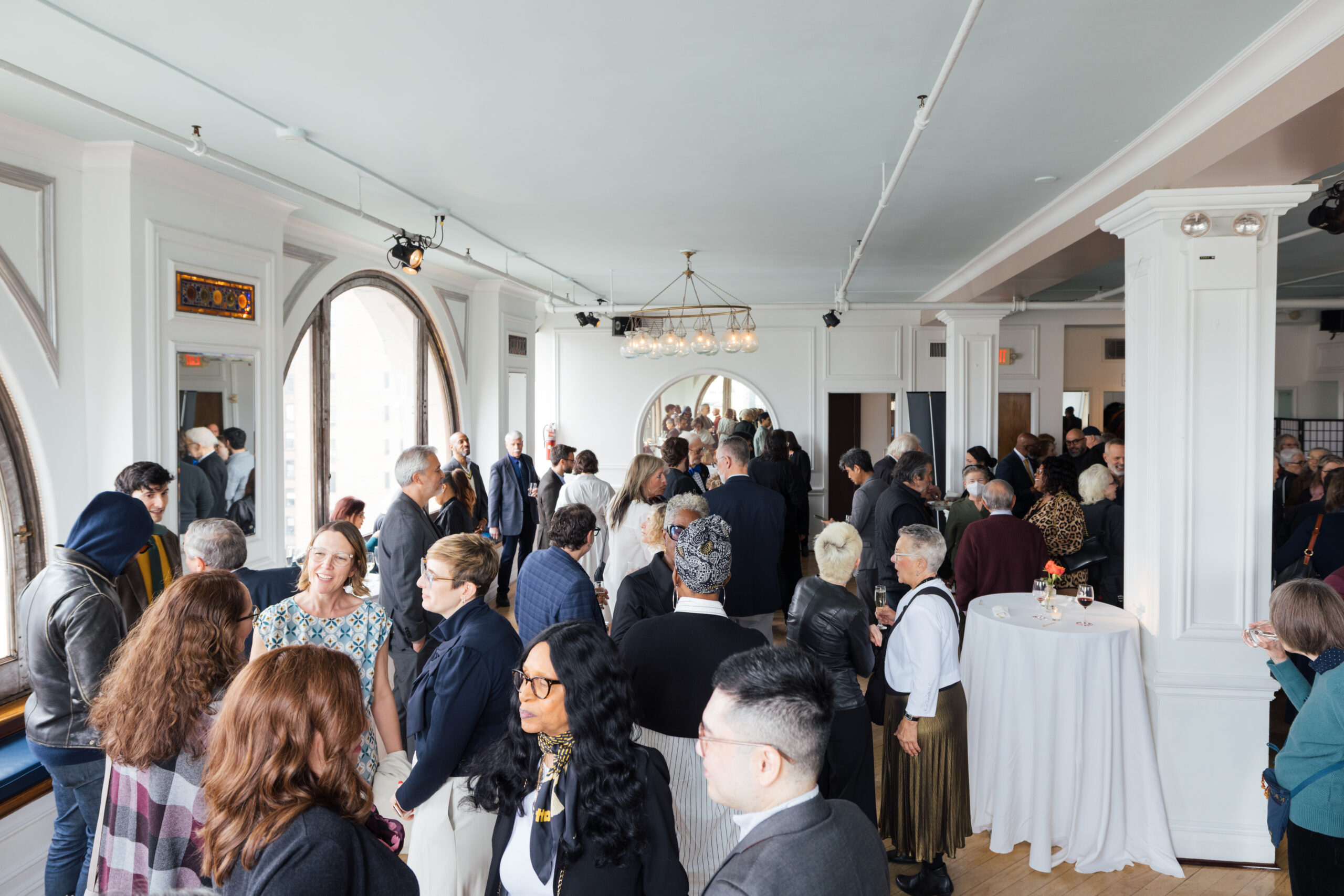 A crowded indoor event is taking place, with numerous people mingling and talking. The room features large windows allowing natural light, elegant lighting fixtures, and white walls. Attendees are dressed in a mix of formal and semi-formal attire, with some holding drinks. A table with a floral arrangement is visible, and reflections can be seen in the mirrors. The atmosphere appears social and lively.