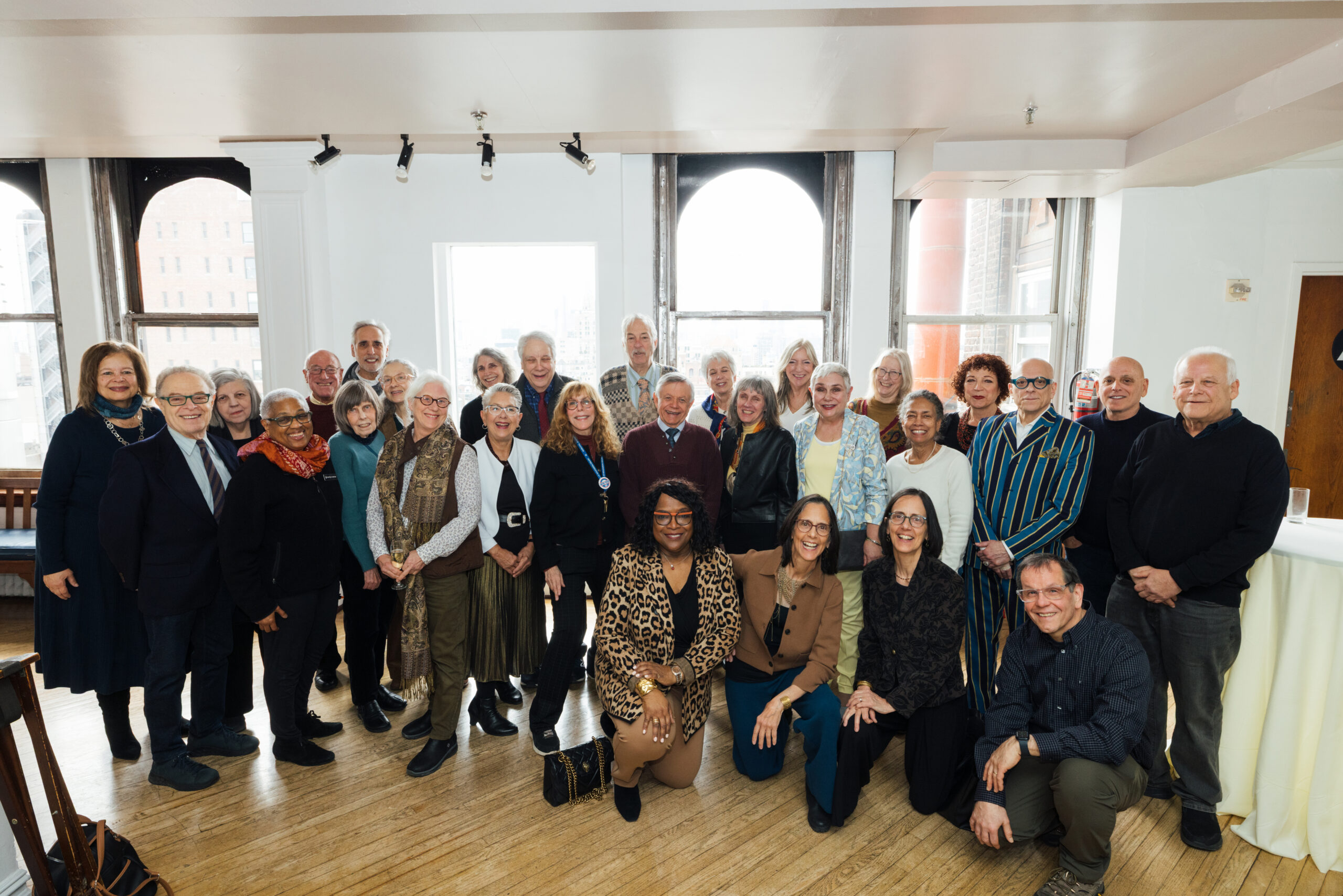 A diverse group of approximately 40 individuals is gathered for a photo in a well-lit room with large windows. They are smiling and posing together in various outfits, showcasing a mix of styles. The background features wood flooring and white walls, with some partially visible furniture on the side.