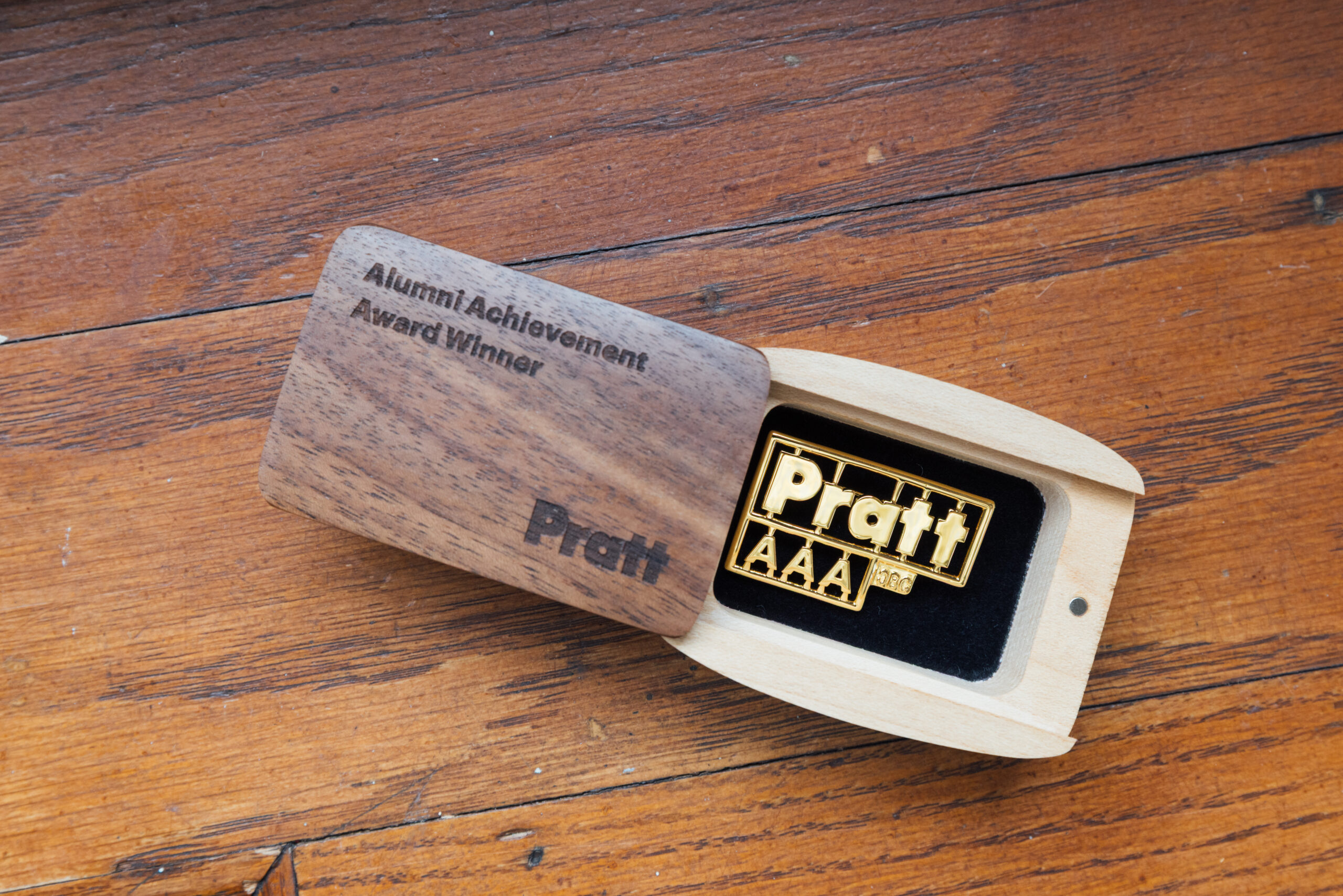 A wooden box is partially open, revealing a gold pin inside that reads "Pratt AAA." The box top features the text "Alumni Achievement Award Winner" engraved on it, with "Pratt" also visible on the side. The background shows a wooden surface.