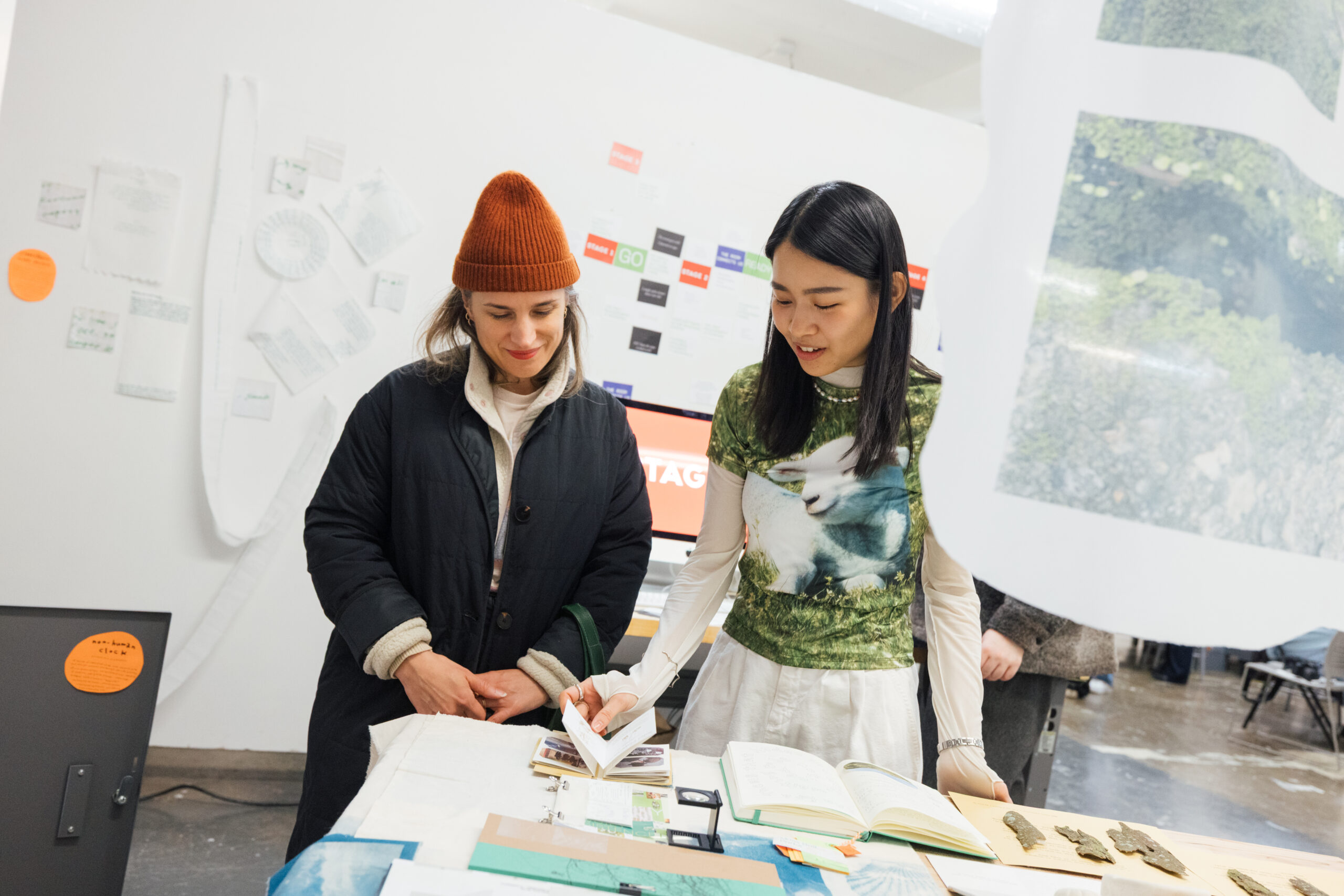 Two women are engaged in discussion at a table filled with various items, including books and printed materials. One woman, wearing a dark coat and an orange beanie, smiles while observing the other, who is wearing a patterned top with an image of a rabbit. In the background, there are posters and other printed materials displayed on the wall. The setting appears to be an art or design exhibition.