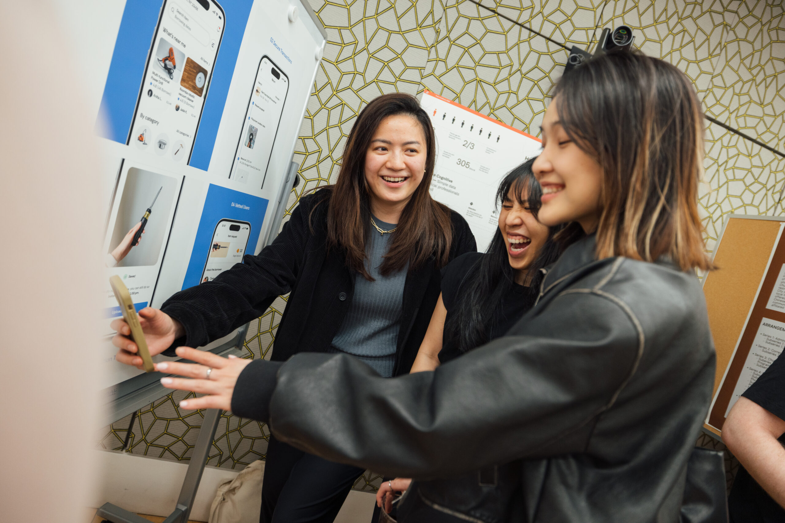 Three women are gathered around a smartphone, smiling and laughing. They appear to be engaged in a discussion about the phone's display, which showcases app designs. Behind them, a wall features a geometric pattern and several informational posters or boards. The atmosphere is lively and collaborative.